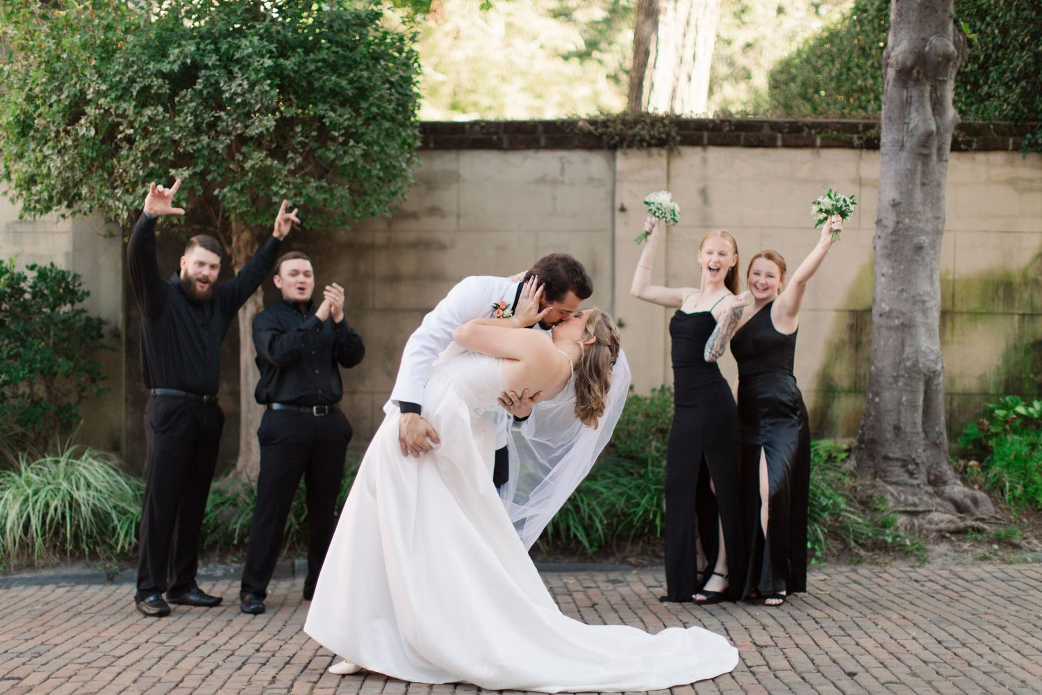 Wedding party members pose joyfully in front of stone pillars and greenery.