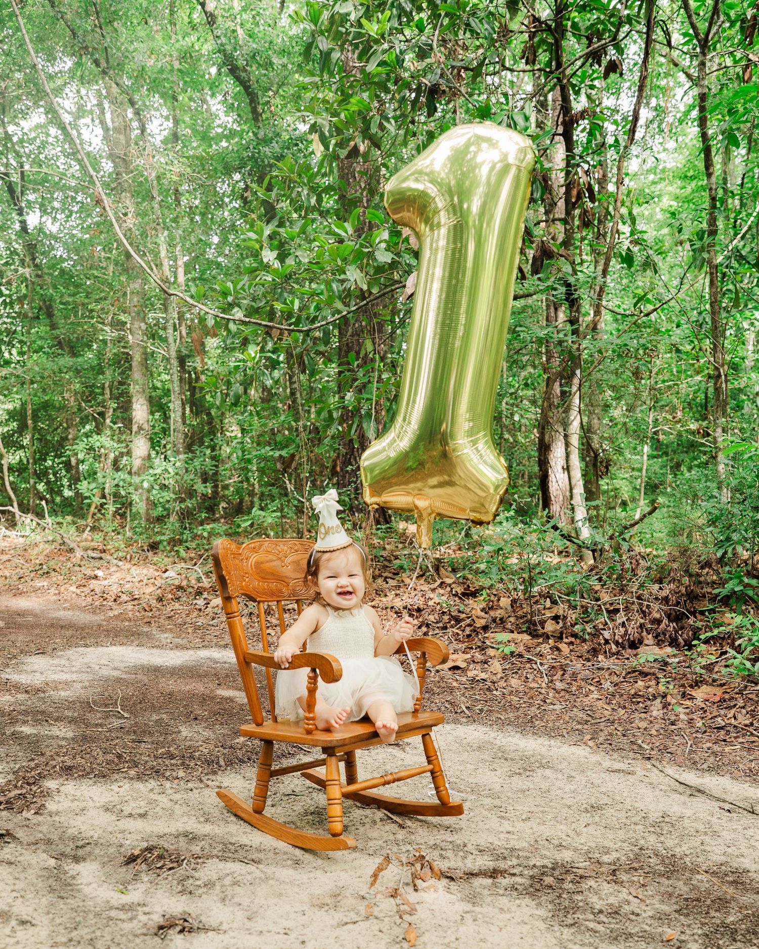 Wooden rocking chair with gold balloon sits on dirt path surrounded by lush green forest.
