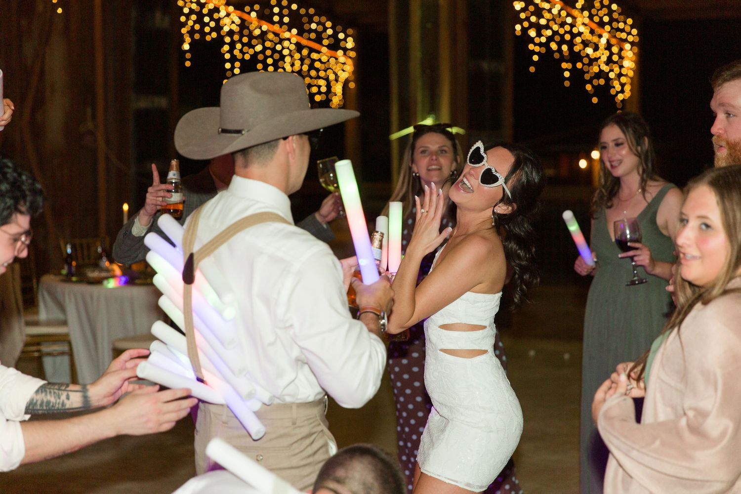 Wedding reception dance party with guests holding glow sticks under ambient lighting.