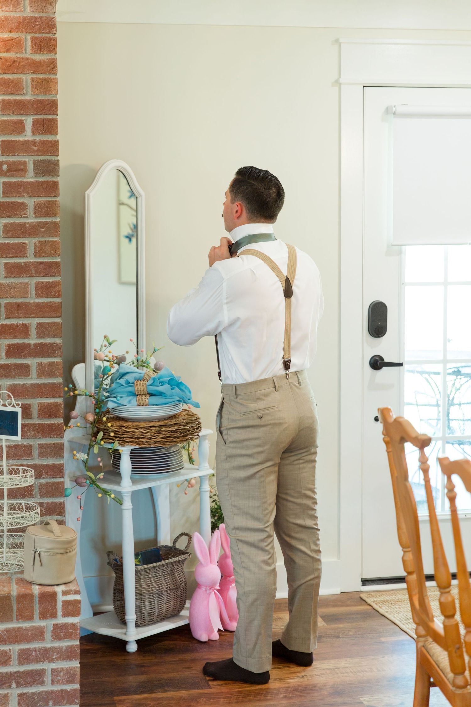 Groom adjusts his tie in front of mirror while getting ready, with vintage decor visible in background.