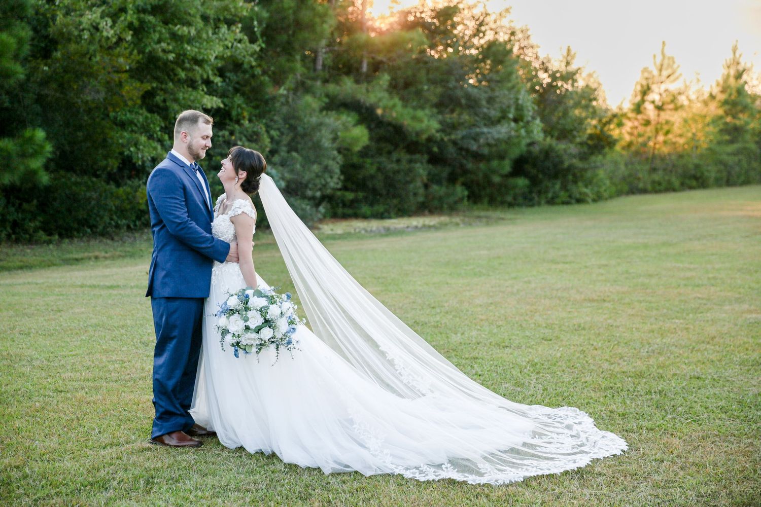 Sunset wedding photos in a grassy field with long bridal veil trailing behind.