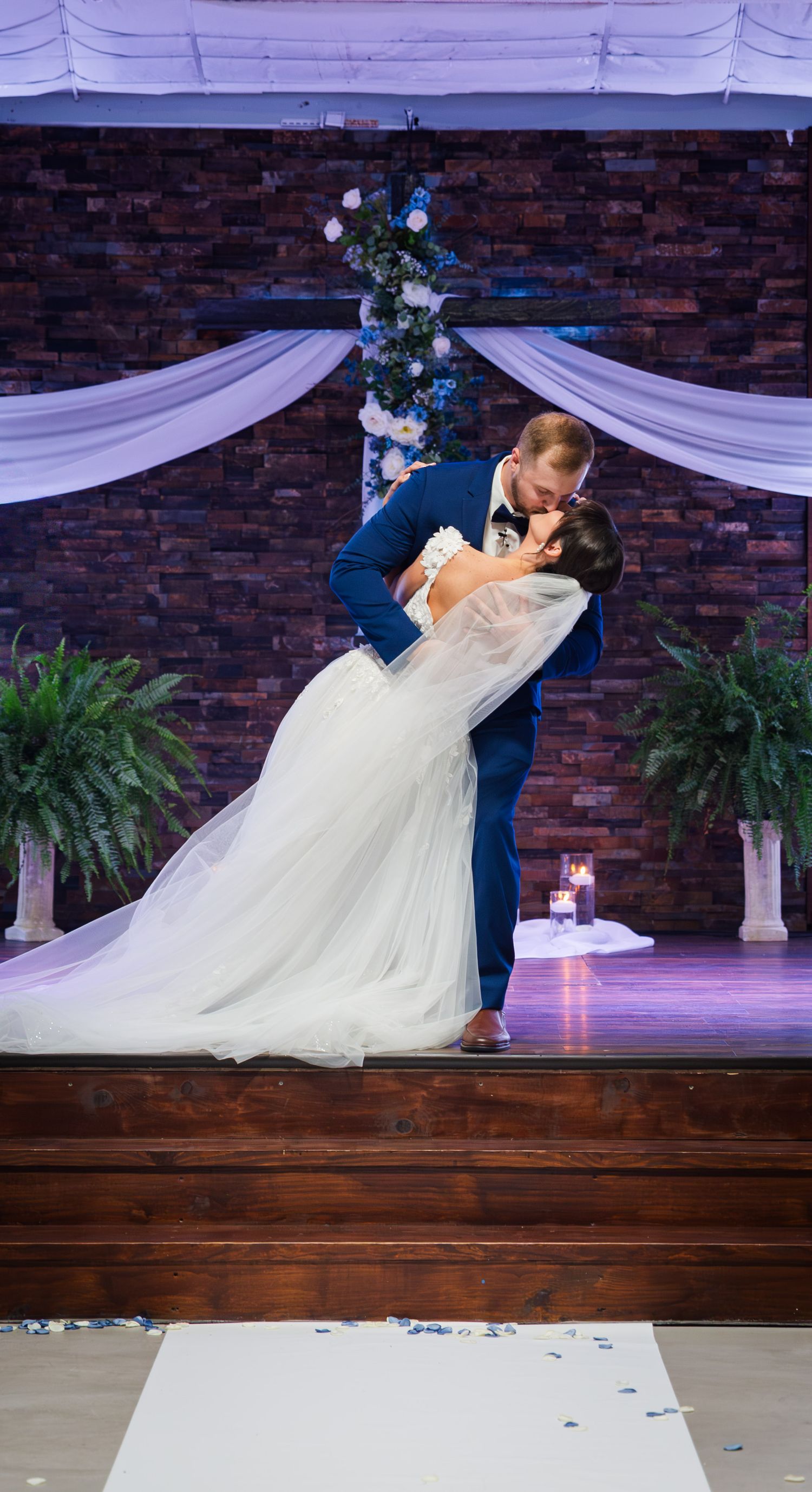 Romantic dip and kiss between newlyweds under draped white fabric and brick wall backdrop.