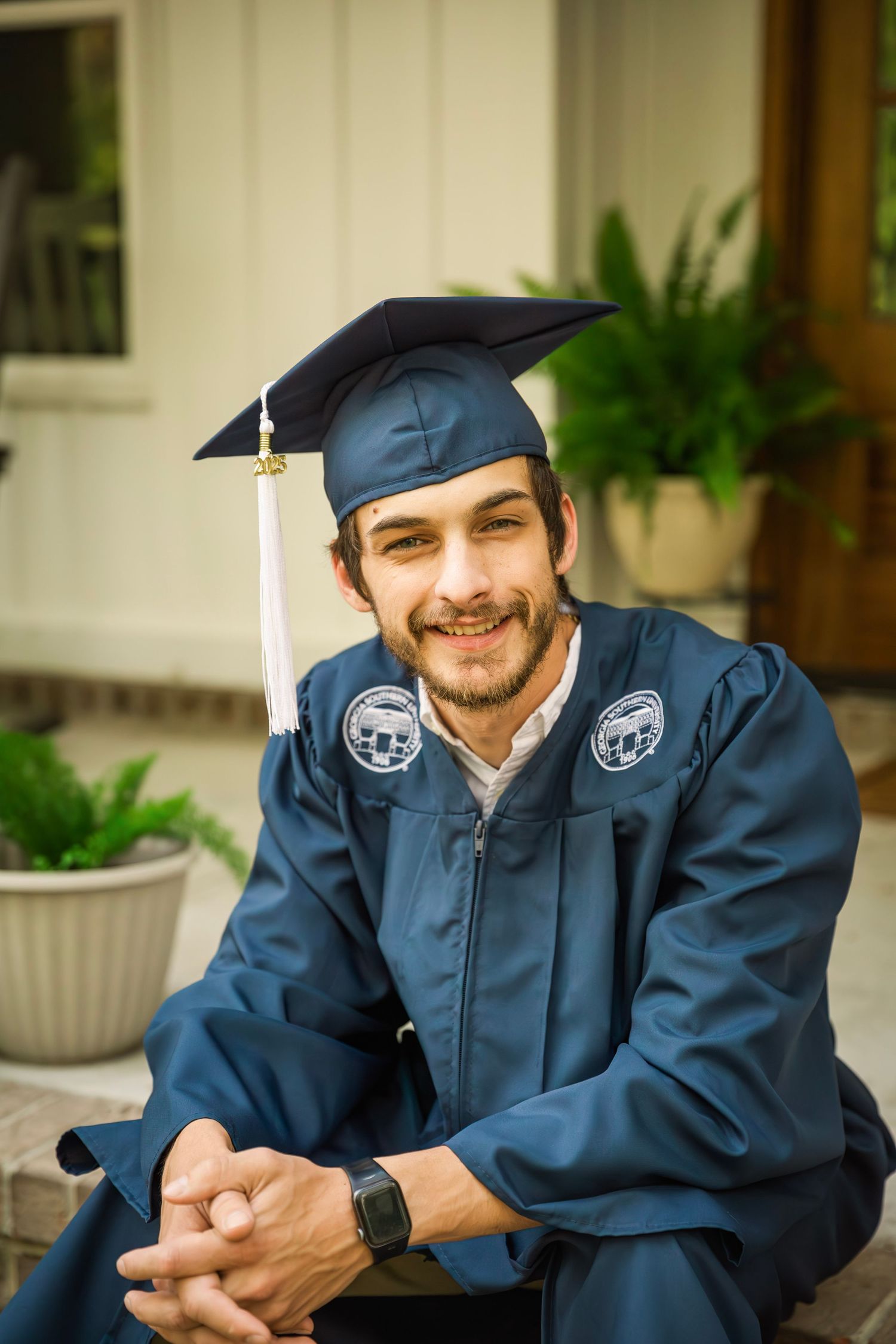 Graduate in navy blue cap and gown sitting on porch steps outdoors.