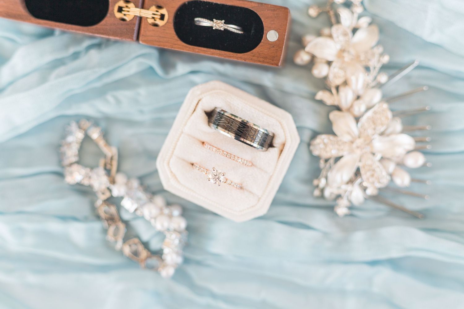 Wedding rings and delicate jewelry displayed on light blue fabric with floral accents.