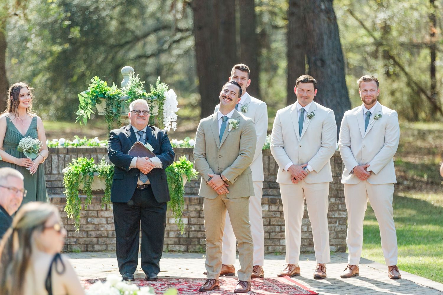 Wedding party members stand in line outdoors wearing coordinated khaki and navy attire while holding bouquets.