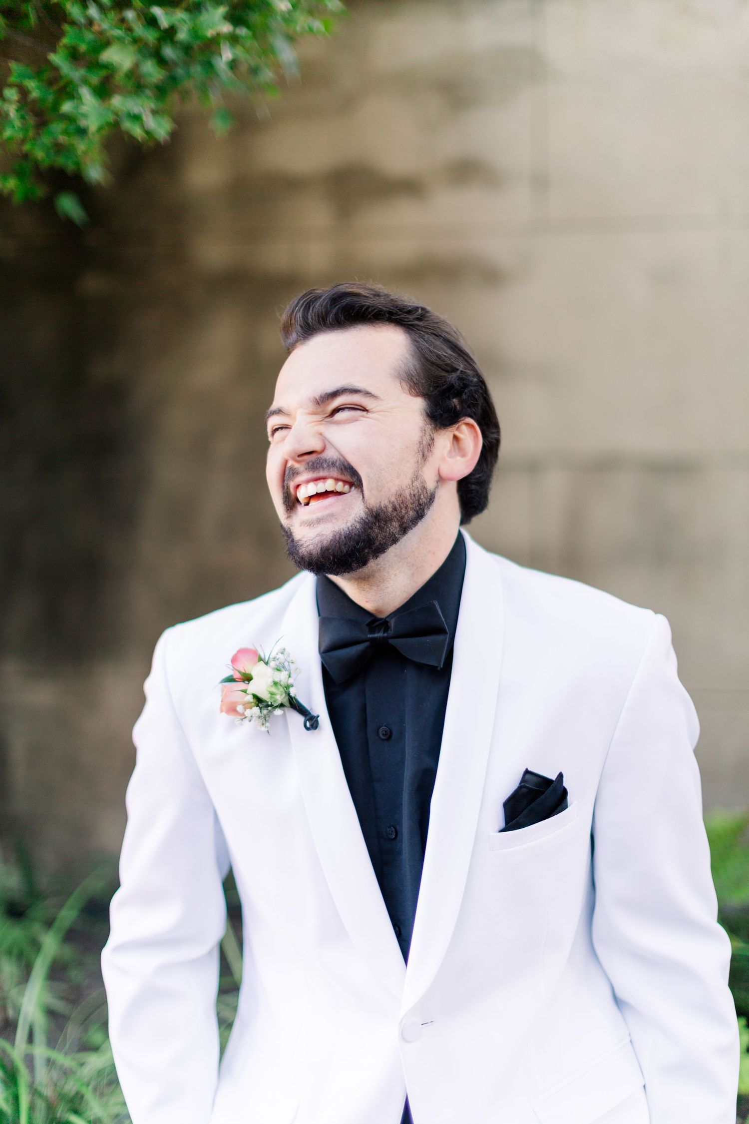 Groom in white jacket and black shirt poses against natural outdoor backdrop.