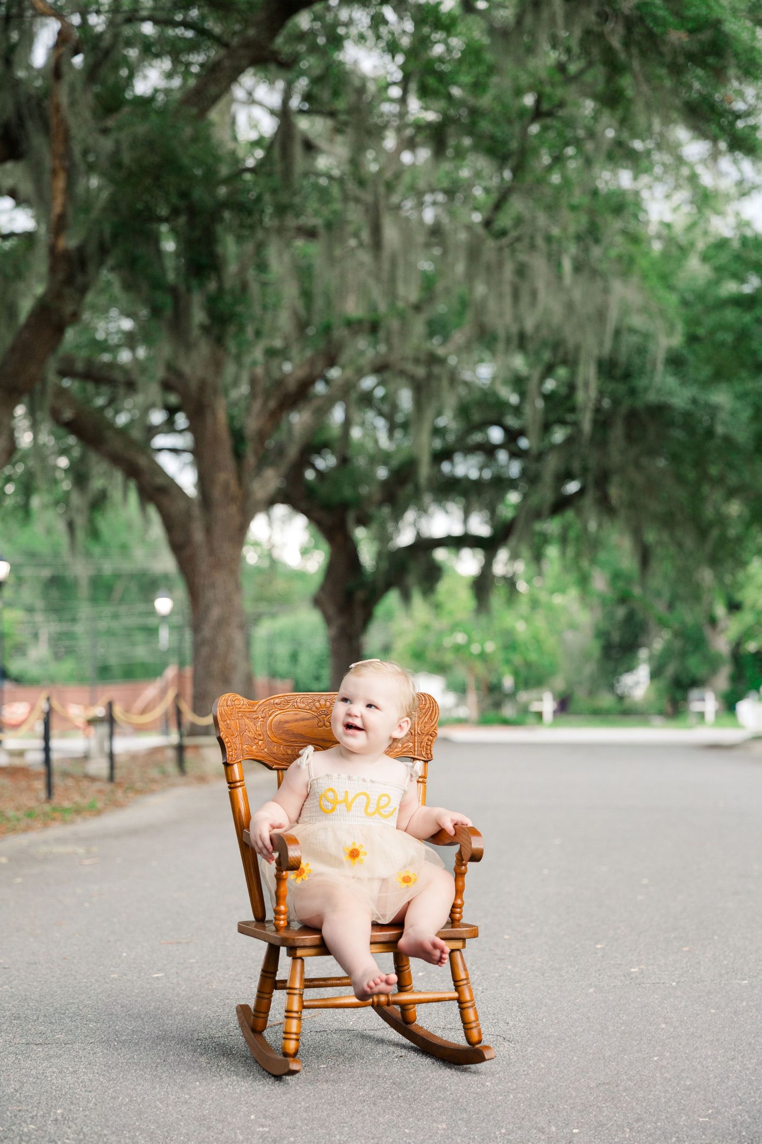 Baby wearing cream dress sits in wooden rocking chair on outdoor path surrounded by trees.
