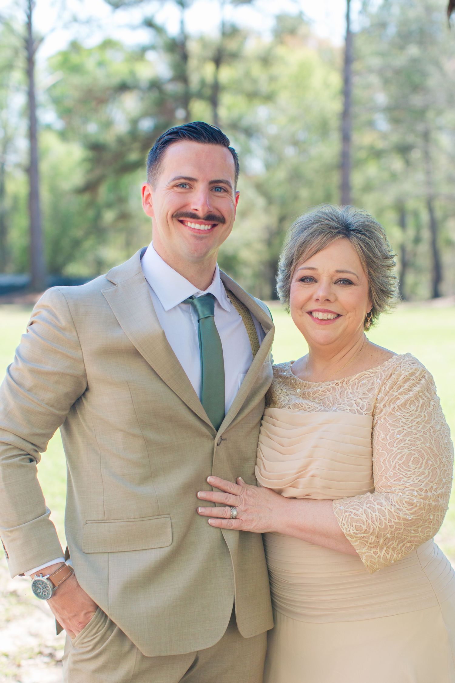 Wedding party in khaki suits and cream dresses pose together outdoors in a pine forest setting.