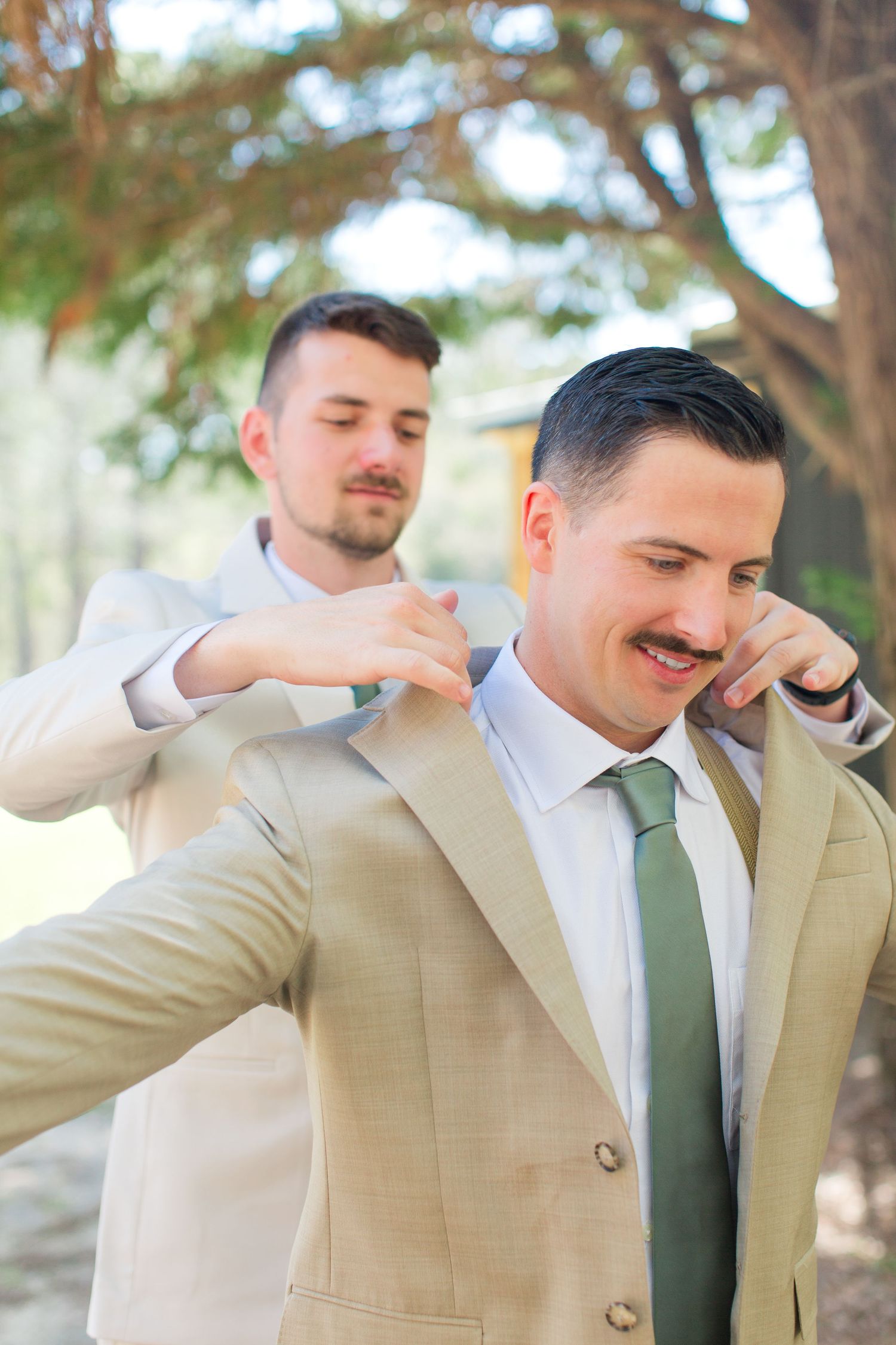 Two groomsmen share a lighthearted moment in tan suits and green ties during outdoor wedding preparations.