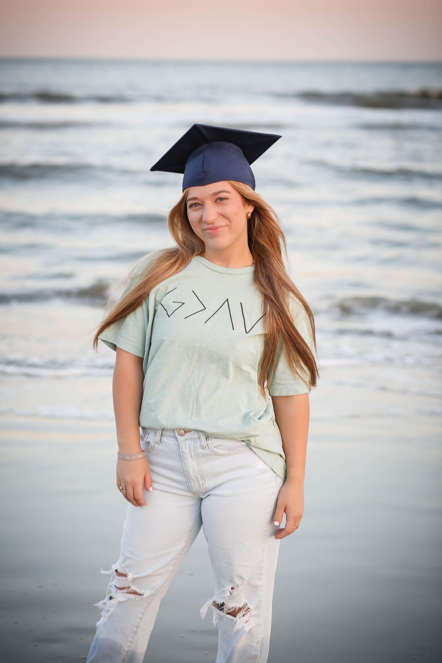 Graduate in casual attire poses on a beach with waves in the background.