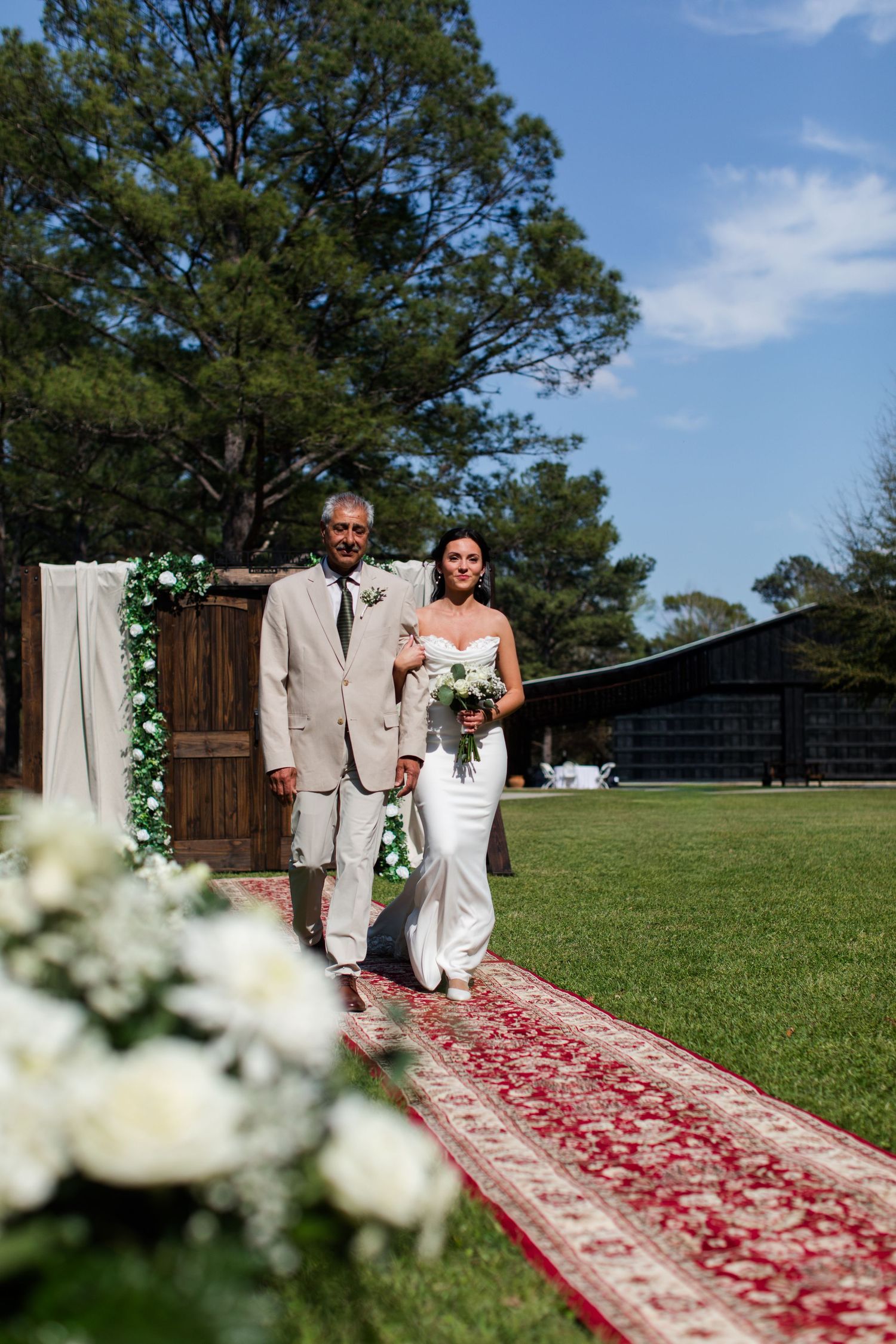 Wedding ceremony processional on an outdoor red carpet aisle with white floral decorations and green lawn backdrop.