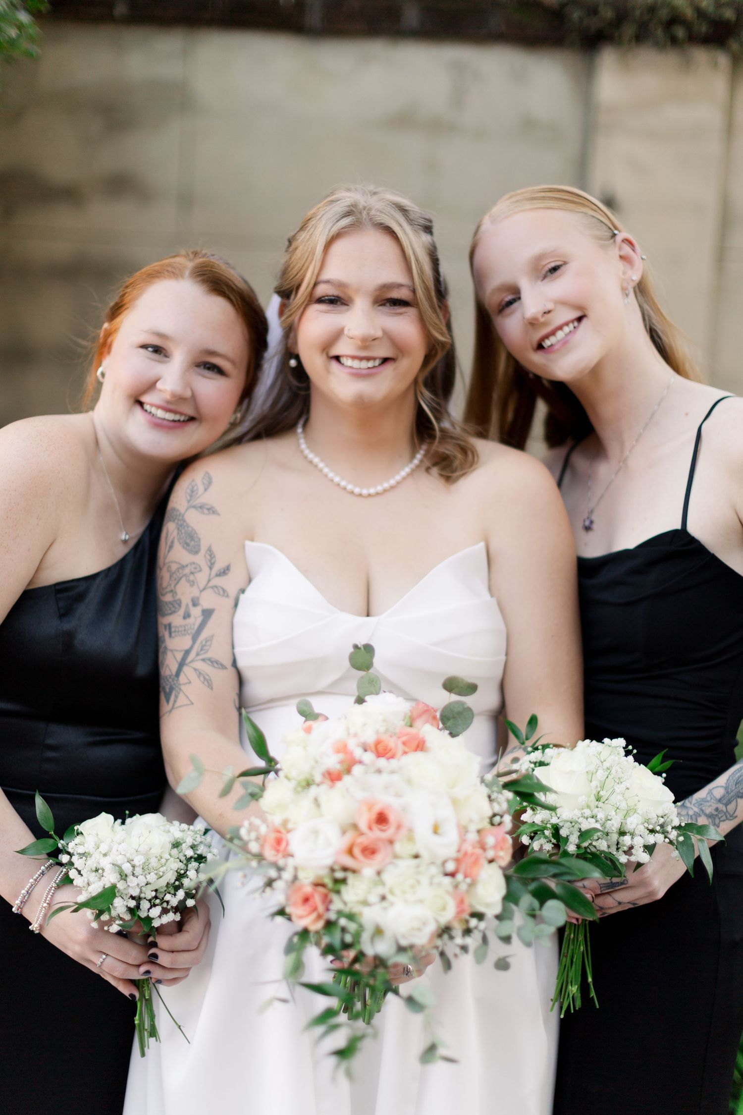 Bridesmaids in black dresses hold coral and white floral bouquets while standing together.
