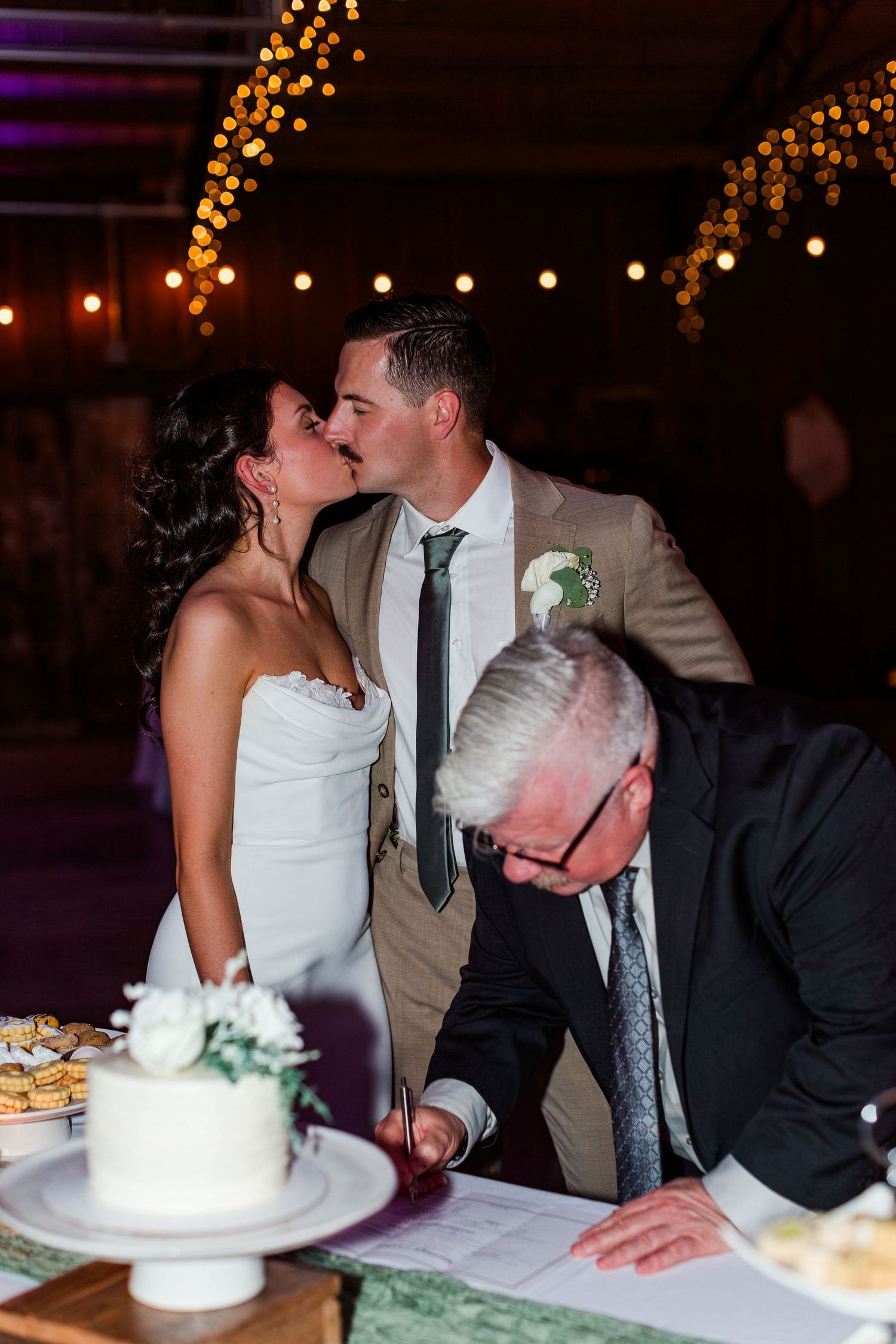 Wedding couple kisses while signing marriage documents beside their elegant white cake under string lights.