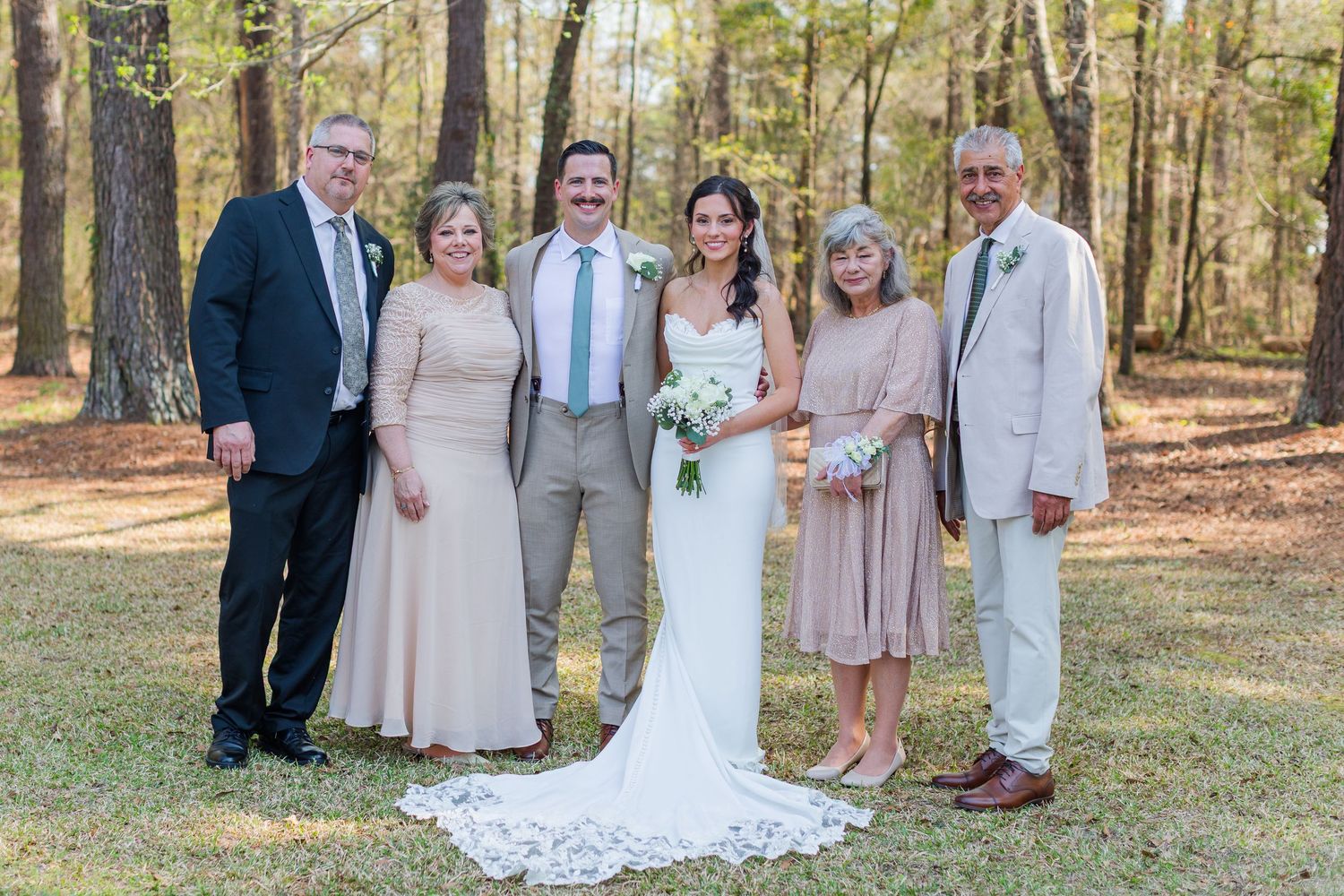 Wedding party in soft pastel colors poses together in a sunlit wooded outdoor setting.