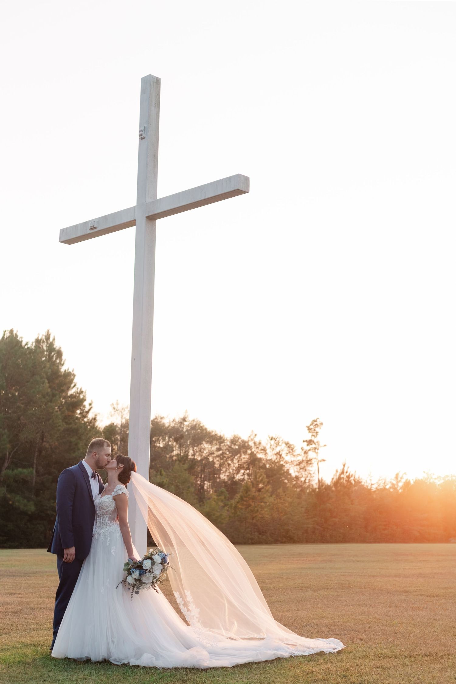 Wedding couple silhouetted against large white cross at sunset.