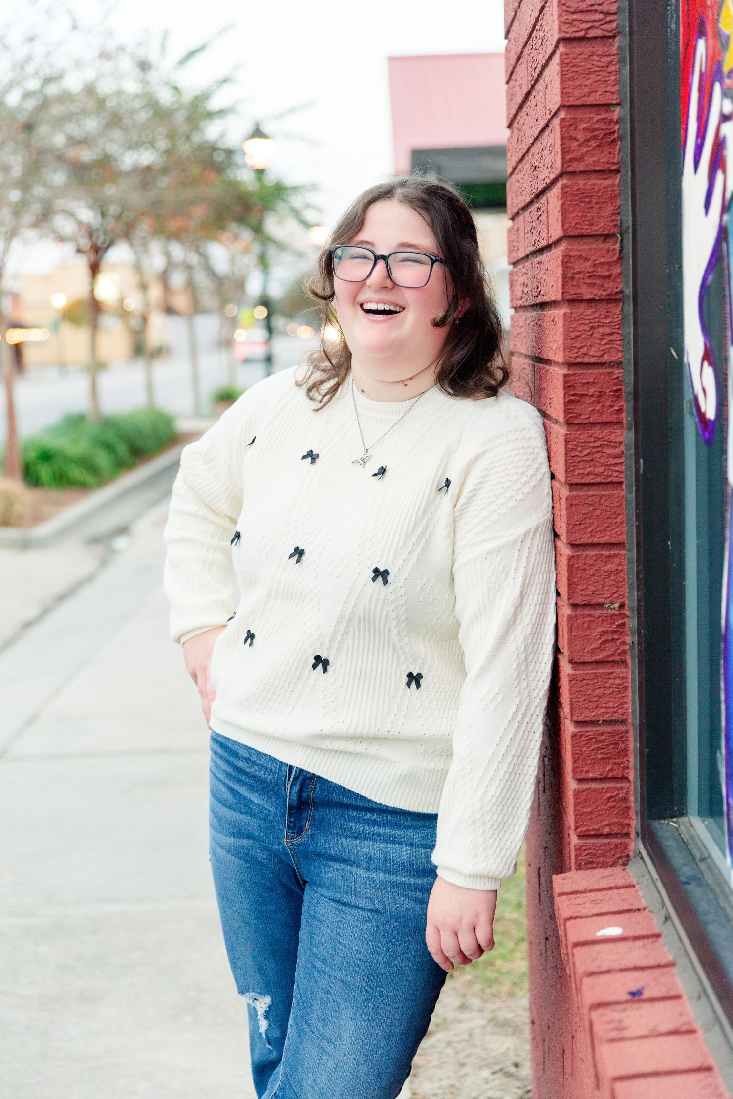 Person wearing white patterned sweater and jeans poses near red brick wall.