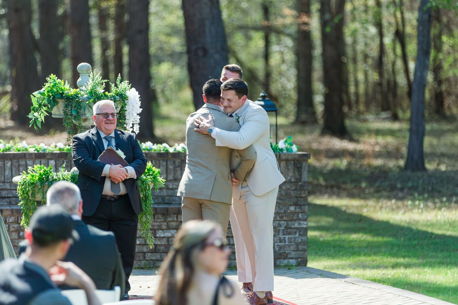 Outdoor wedding ceremony in a sunlit forest setting with guests seated on wooden benches.