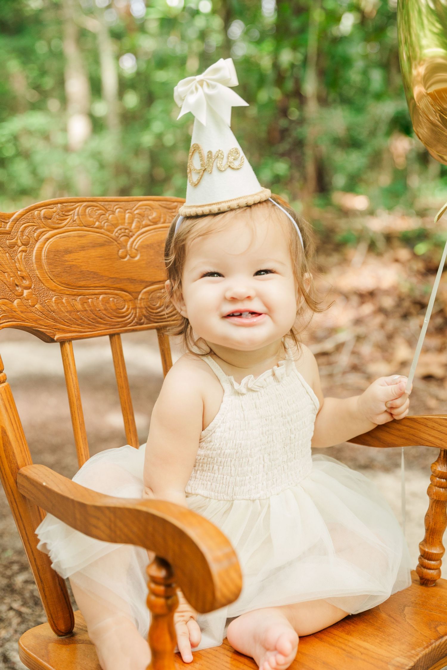 Baby in white dress and party hat sitting on wooden vintage chair.