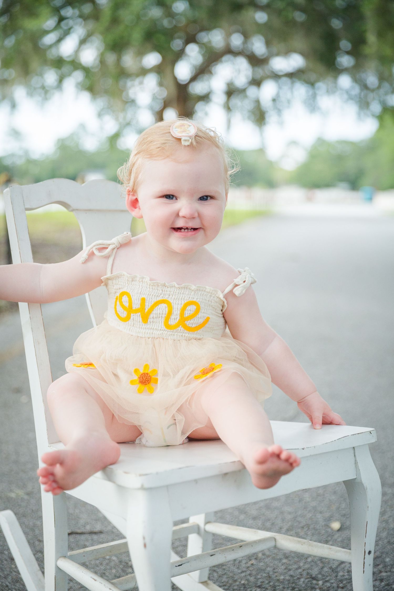 Baby in yellow dress sits on white bench outdoors with joyful expression.