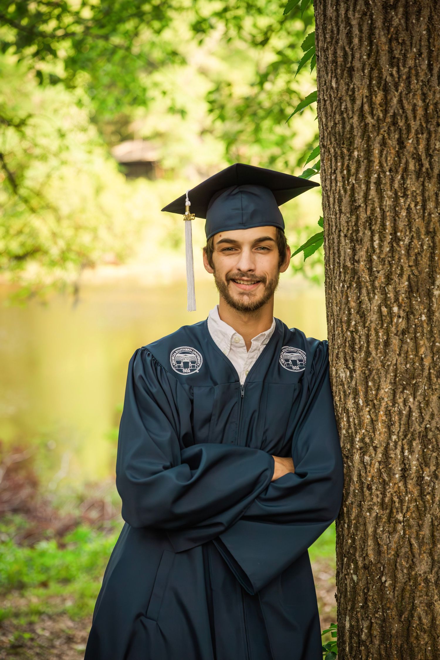 Graduate in black cap and gown posing between trees in outdoor setting.