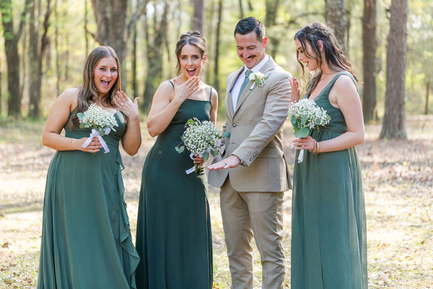 Wedding party in emerald green dresses and grey suits gather outdoors in a sunlit wooded setting.