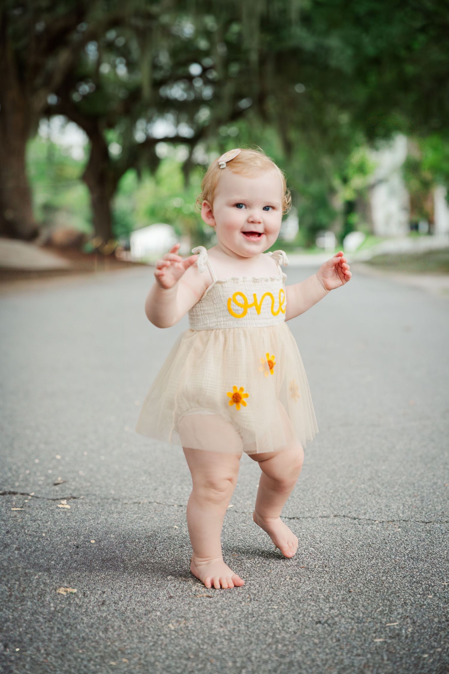 A baby in a peach tulle outfit takes first steps on pavement outdoors.