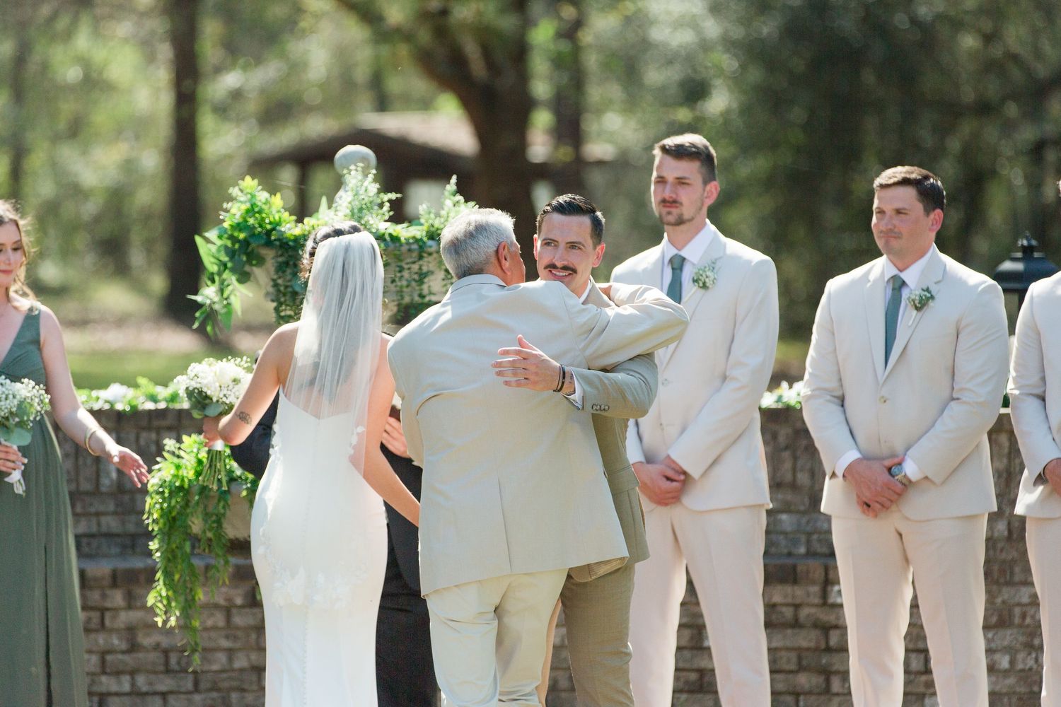 Wedding party in light colored attire stands in line against brick wall and greenery outdoors.