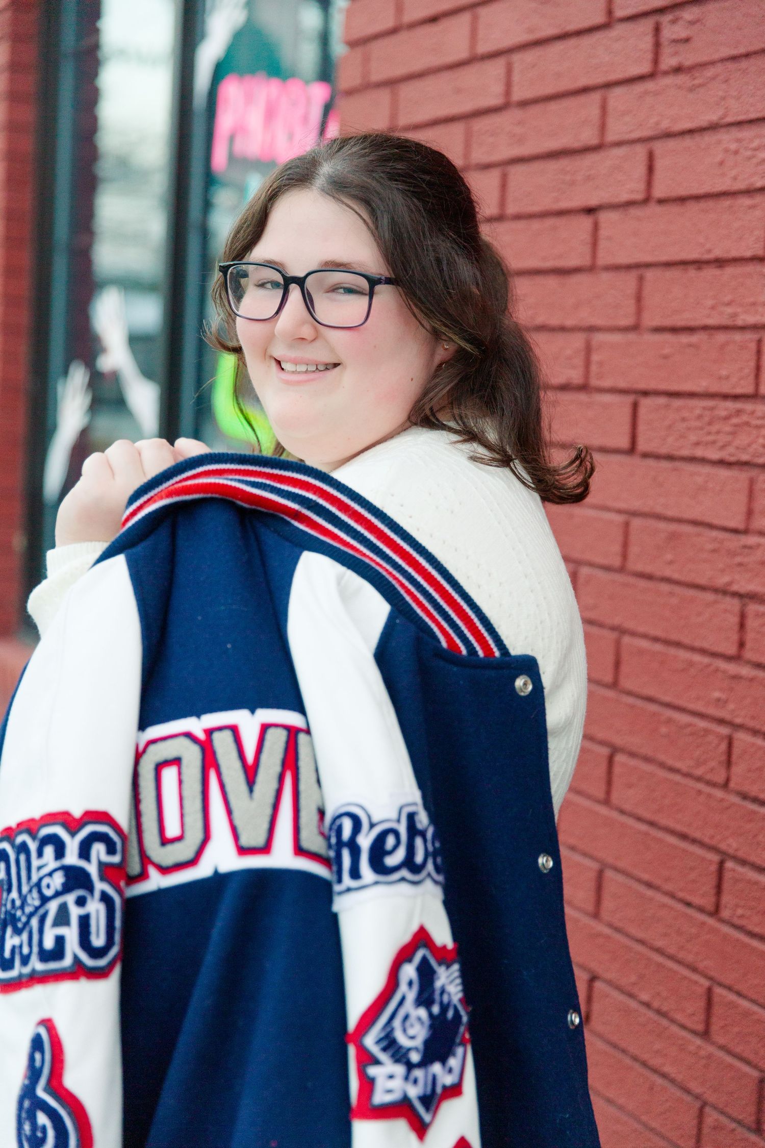 Student wearing varsity letterman jacket with white and blue colors against red brick wall.