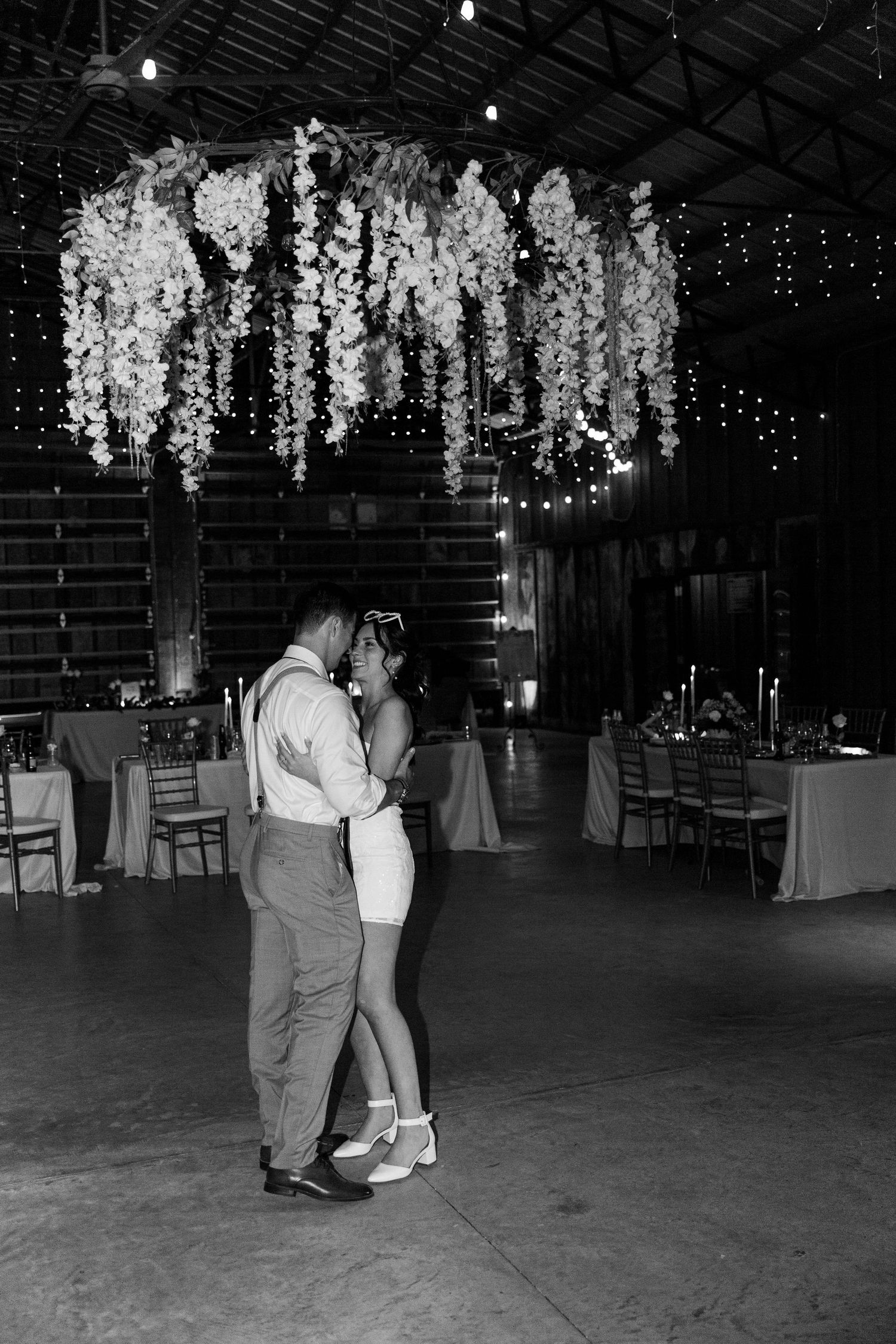 Couples dancing under hanging white floral decorations and string lights in a dimly lit reception venue.