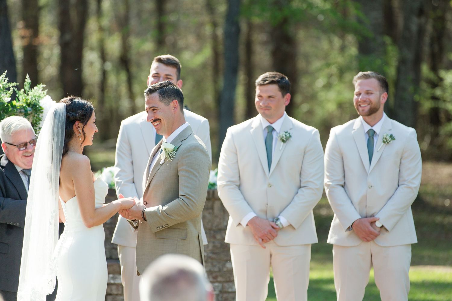Wedding ceremony in progress with bridal party in light suits standing at outdoor forest altar.