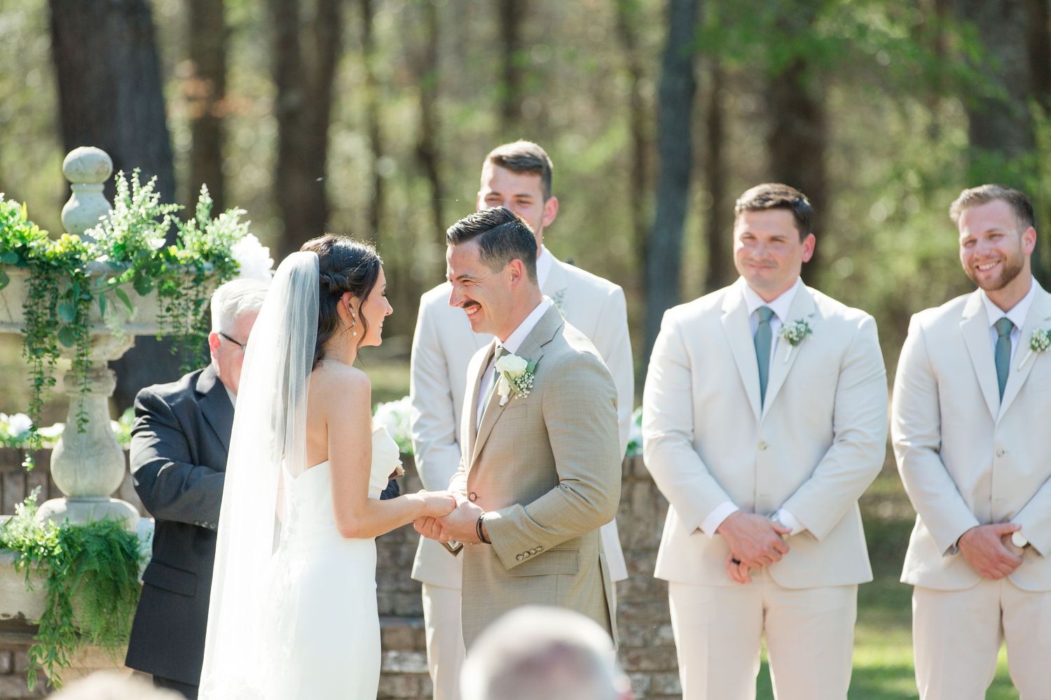 Outdoor forest wedding ceremony with wedding party in light colored suits and floral decorations.