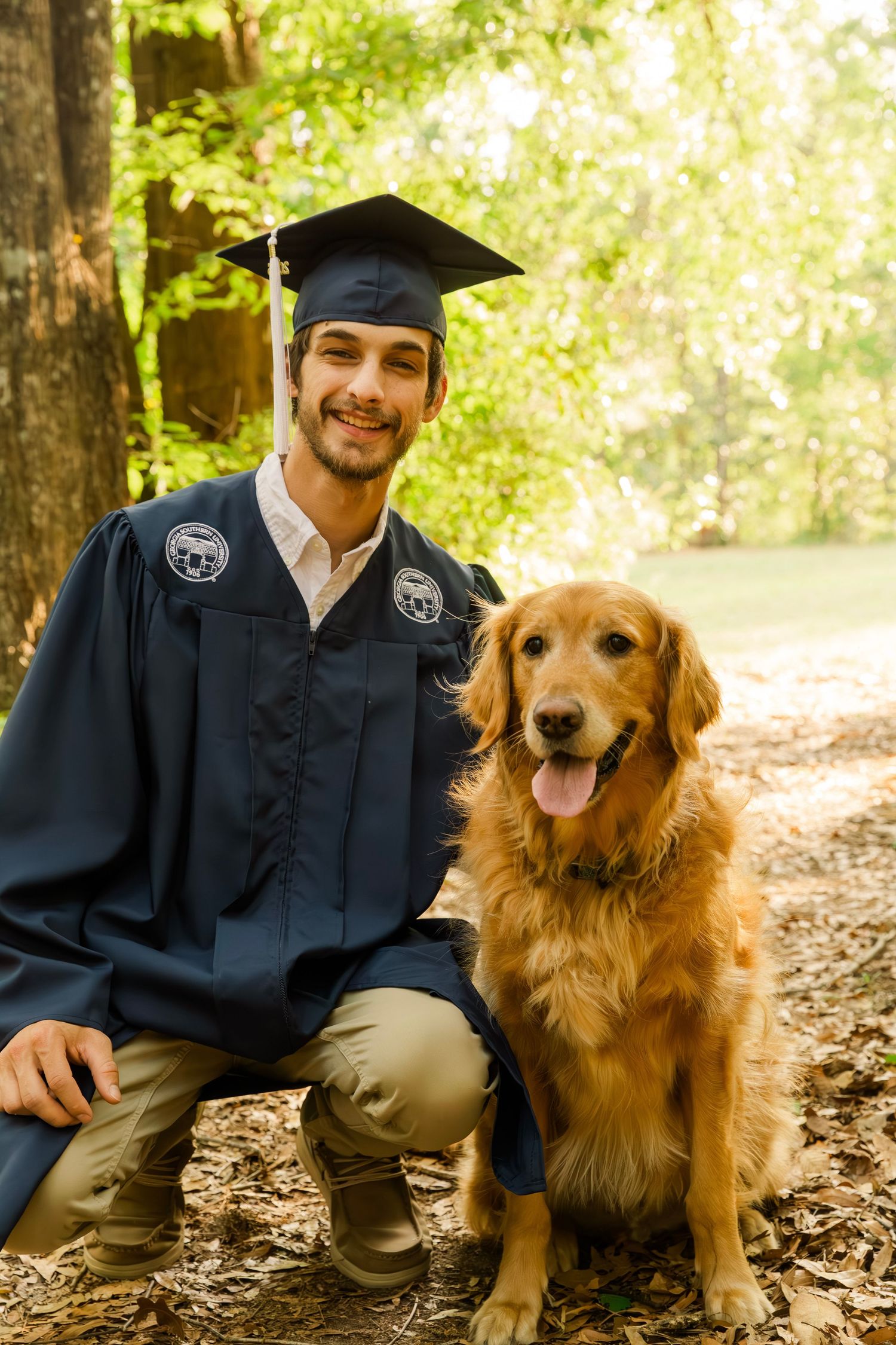 Graduate in black cap and gown kneeling next to golden retriever dog in wooded area.
