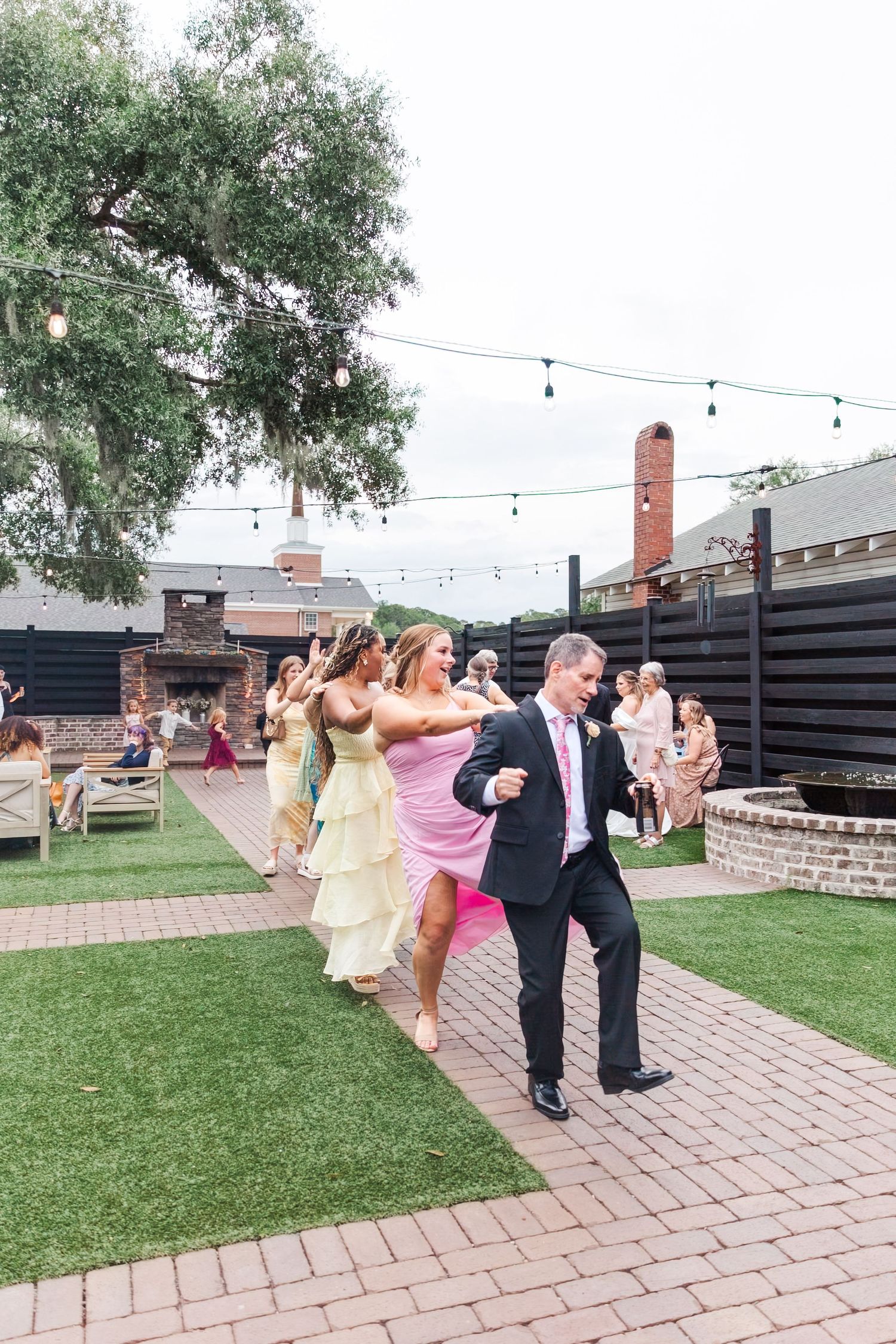 Wedding guests dancing in line on outdoor brick patio with string lights overhead.