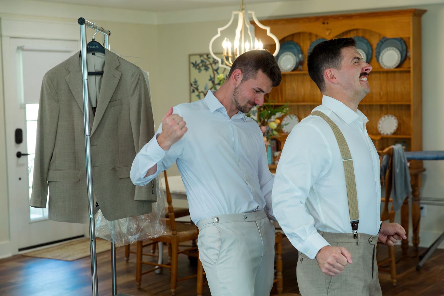 Two people helping each other get ready in formal attire with suspenders and dress shirts in a bright room.