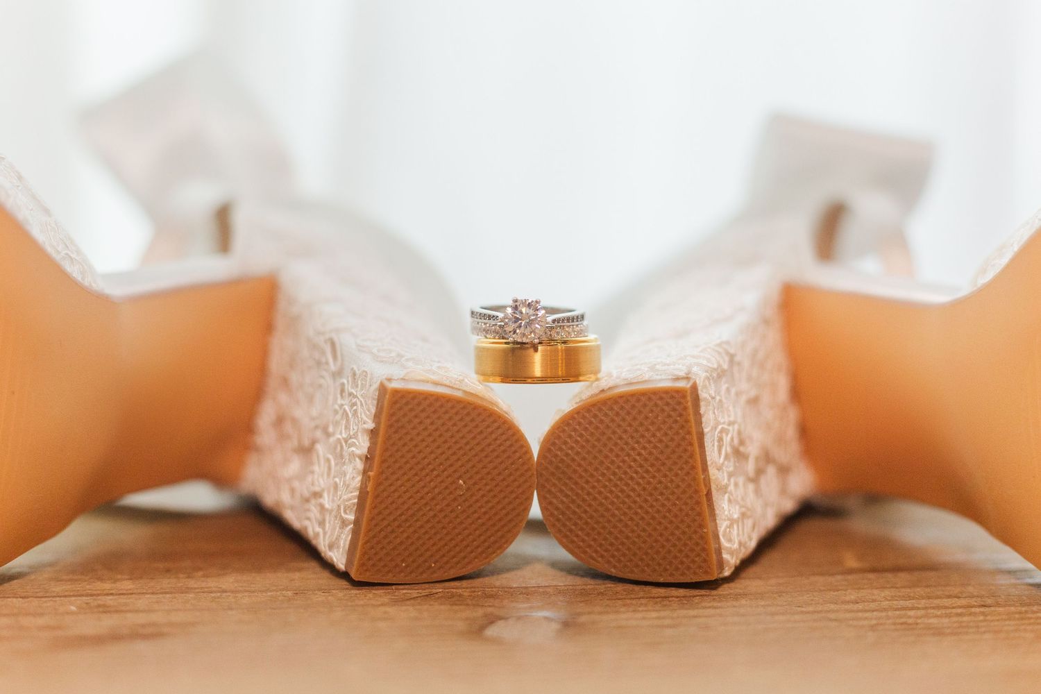 Wedding rings placed artistically between white bridal shoes on wooden surface.