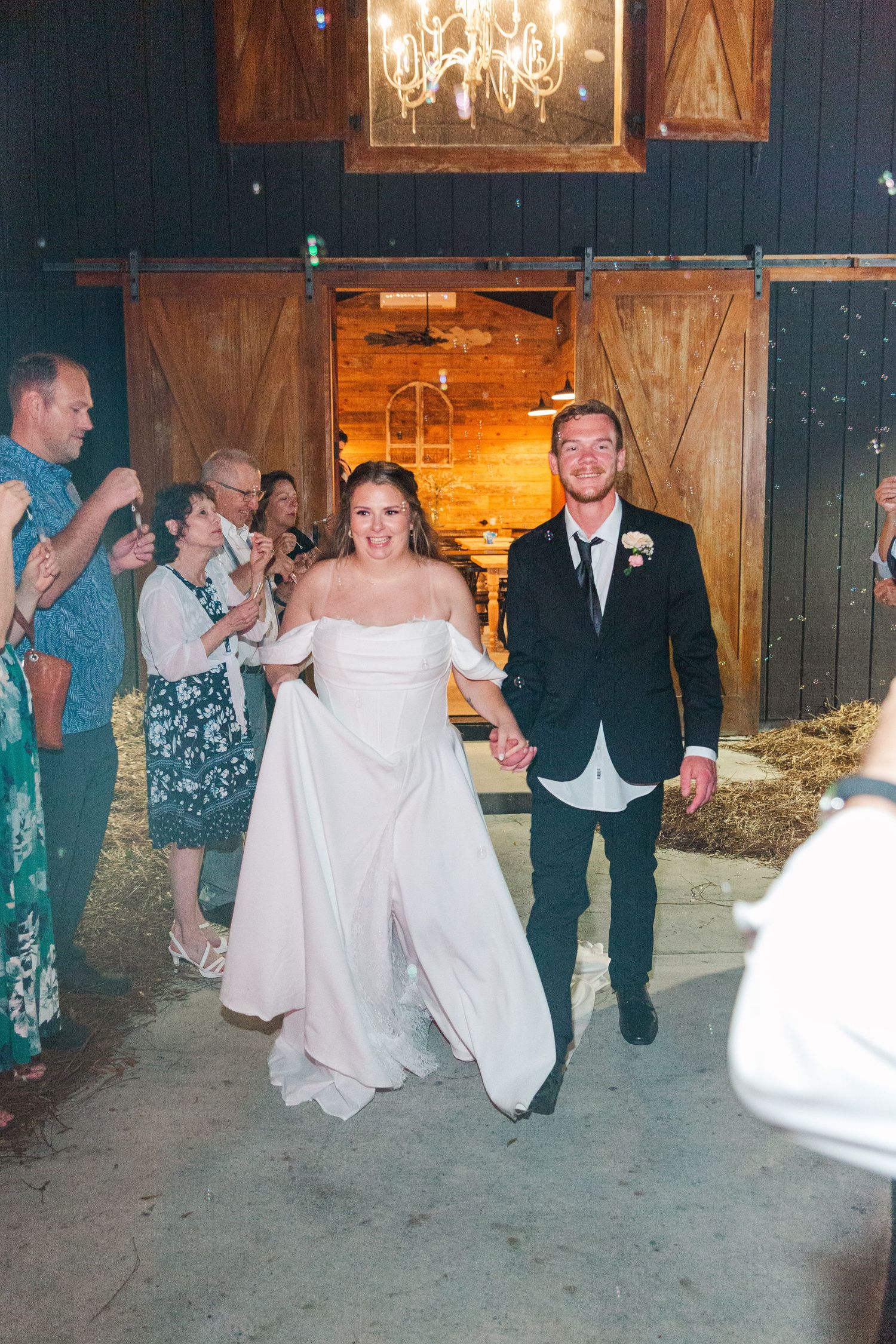 A joyful wedding couple exits their rustic barn venue under a crystal chandelier.
