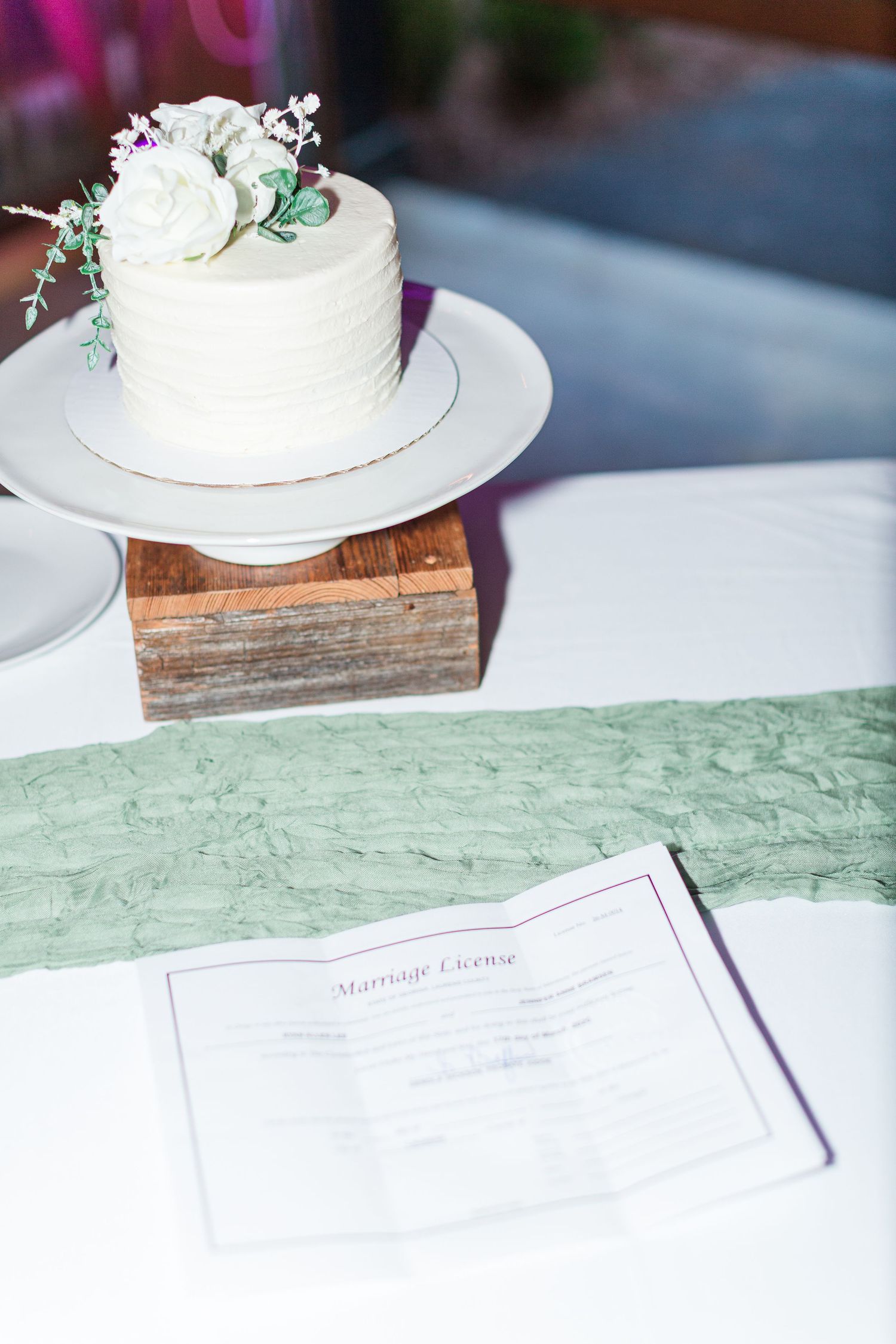 White wedding cake with floral decorations displayed on a wooden stand next to a marriage license.