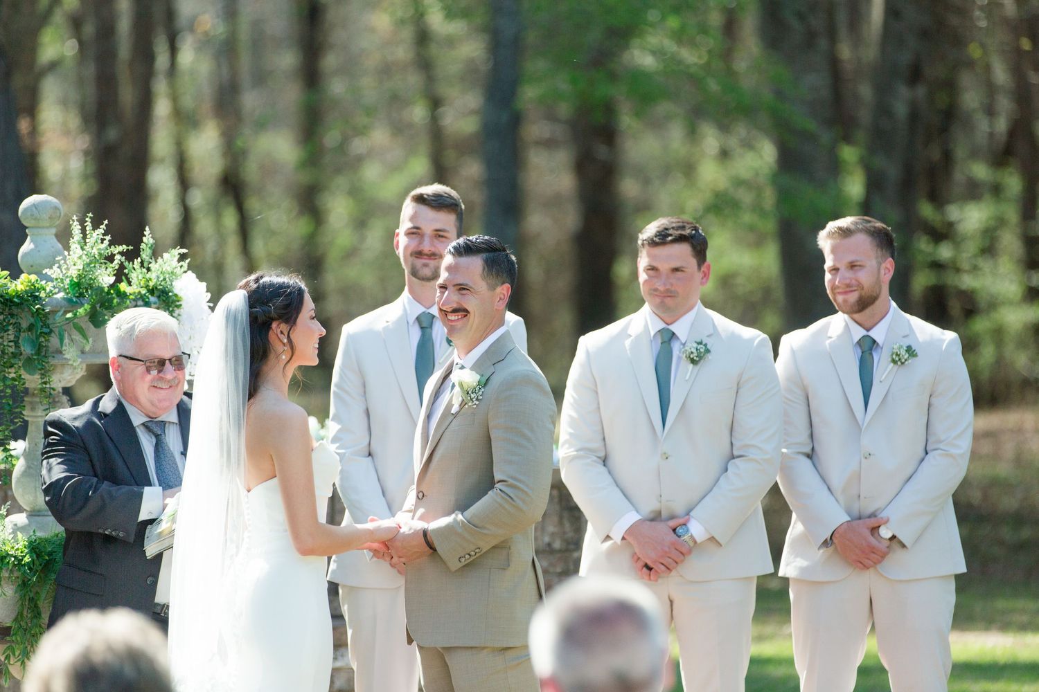 Wedding party members in light colored suits stand together during an outdoor forest ceremony.