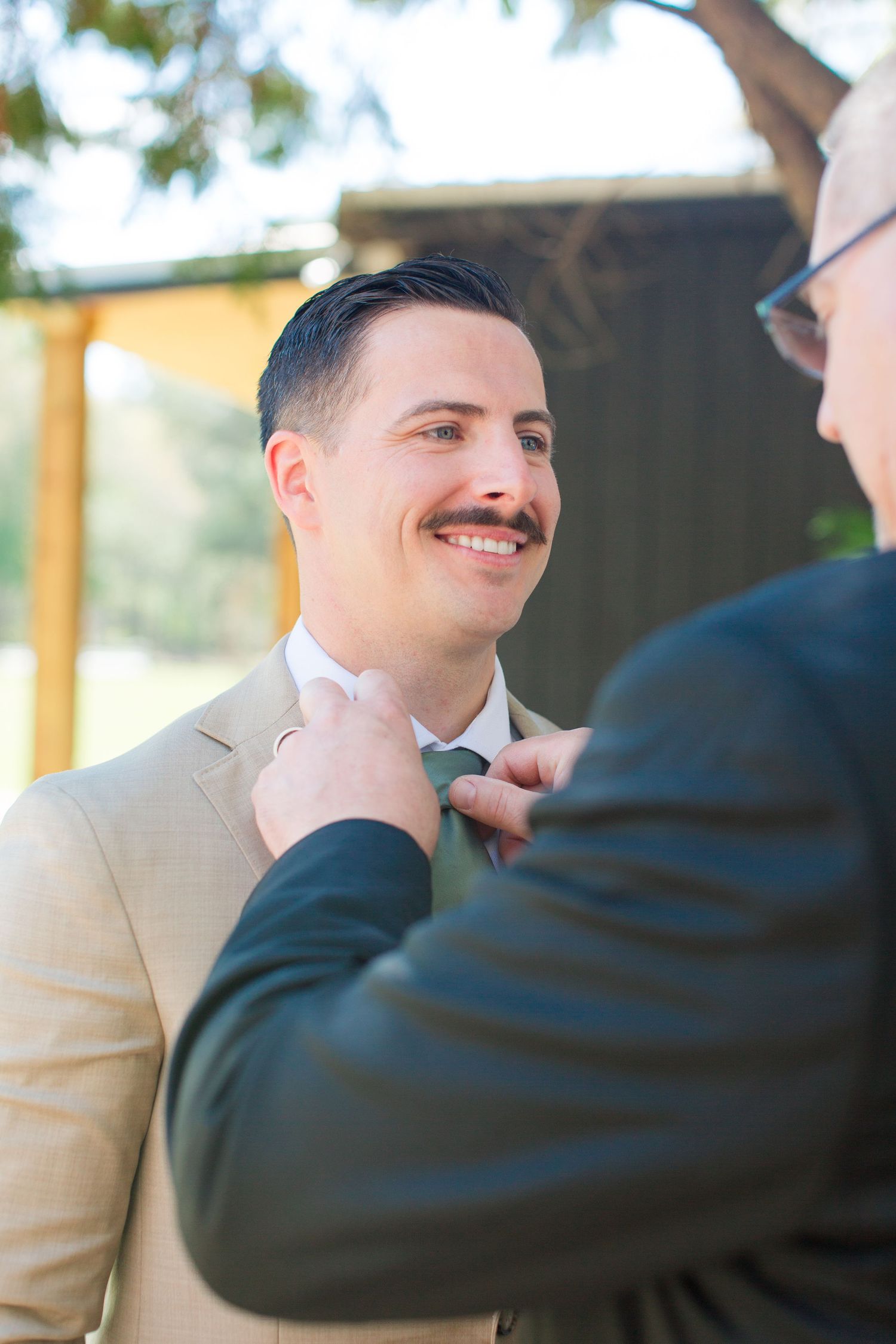Someone adjusts a teal tie on a person wearing a beige suit jacket during what appears to be a wedding preparation.