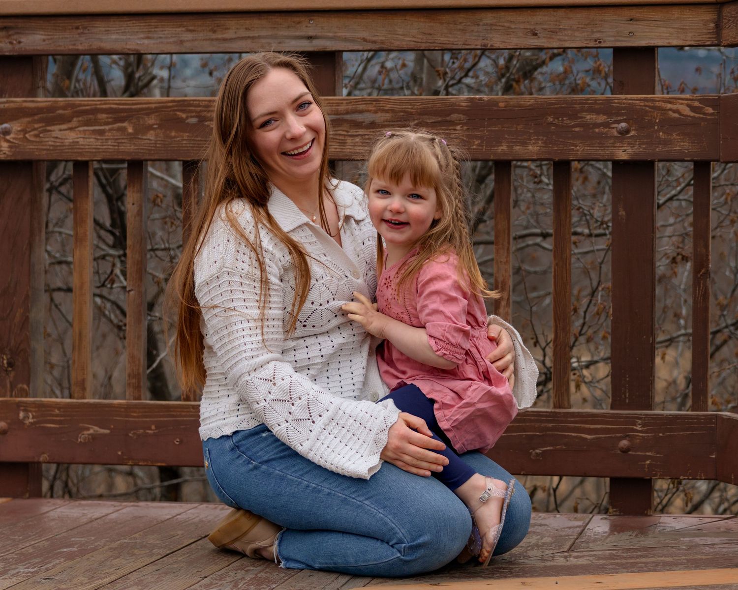 A smiling young woman and little girl pose together on a wooden bridge deck outdoors.