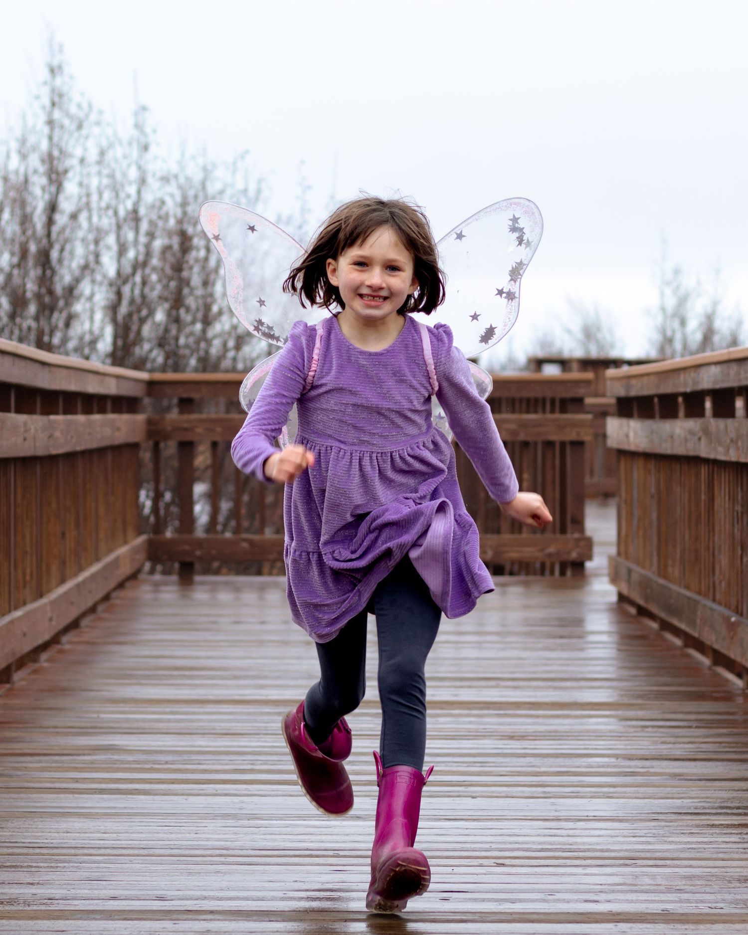 A joyful girl in a purple dress and fairy wings runs on a wooden bridge wearing pink rain boots.