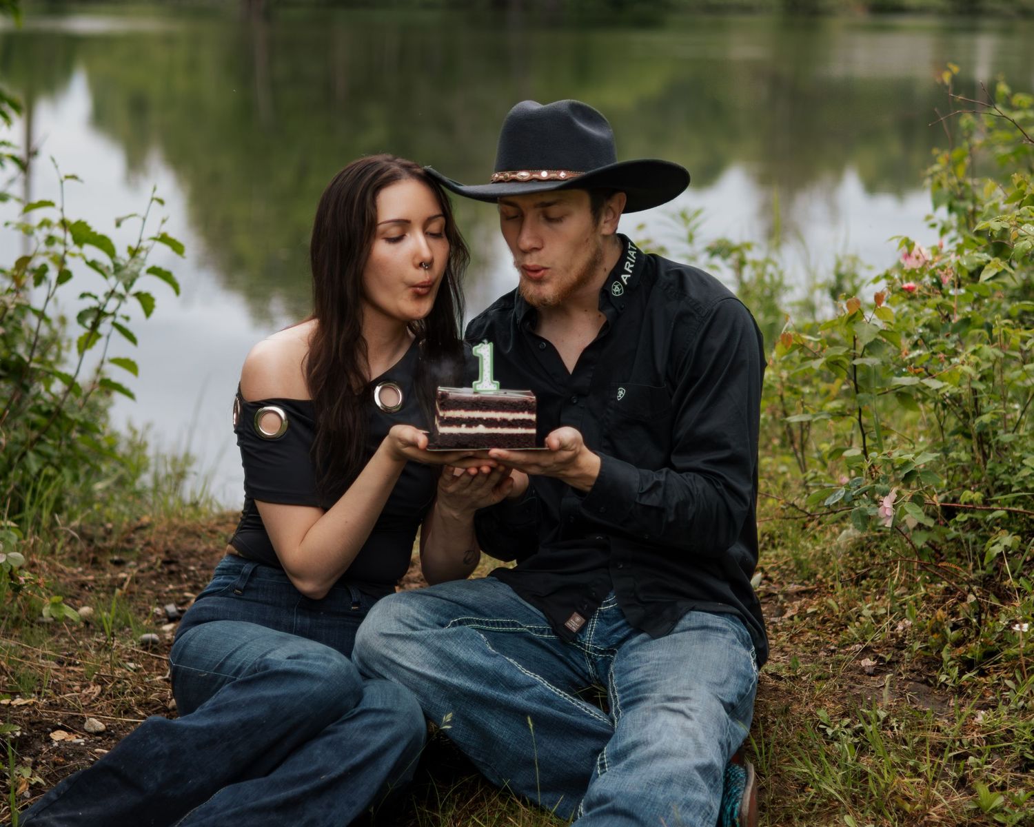 Couple blowing out number one candle on small chocolate cake by a lake, man wearing black cowboy hat.