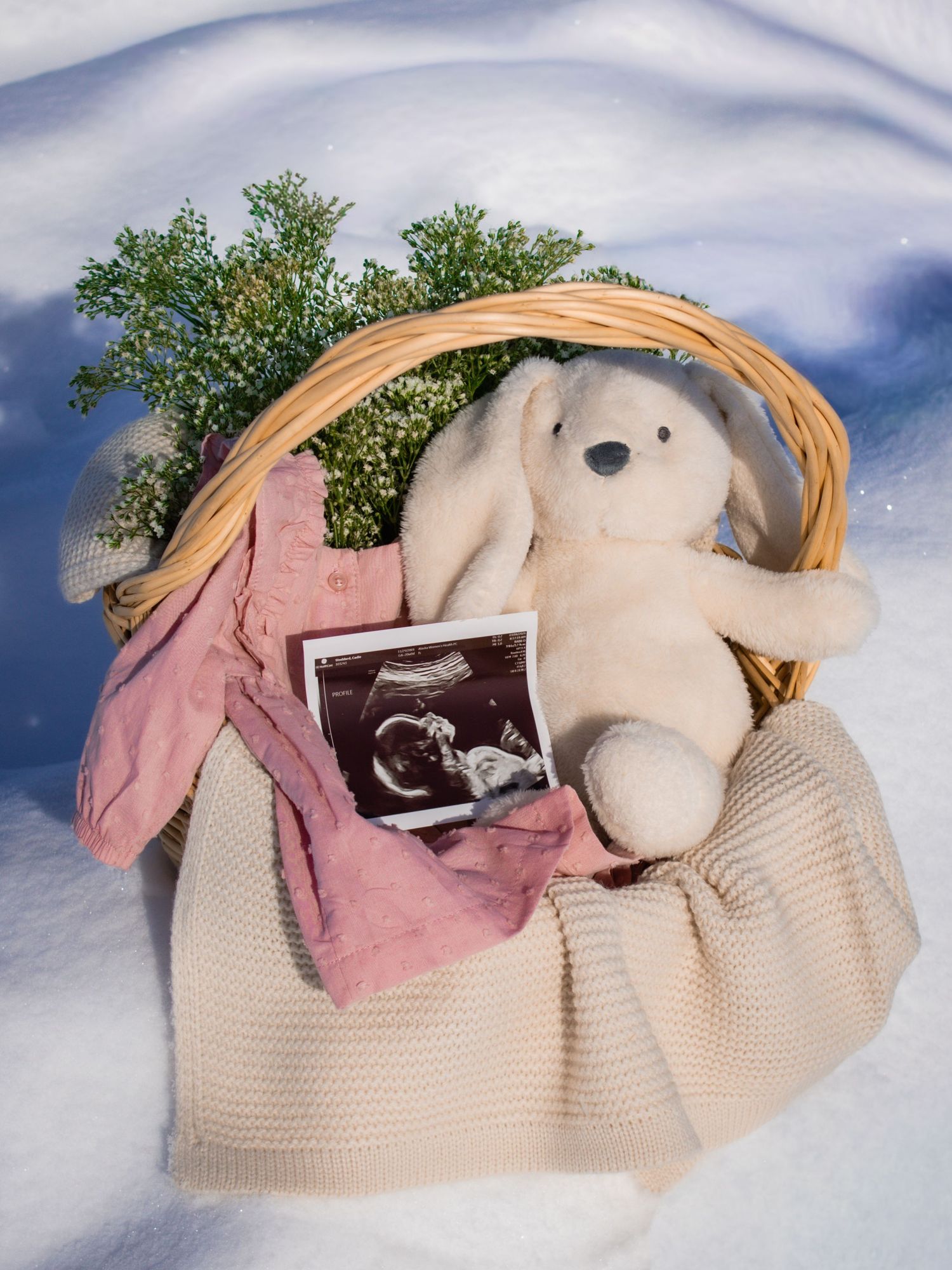 Wicker basket in snow with ultrasound photo, white bunny plush, pink baby clothes, and greenery for pregnancy announcement.