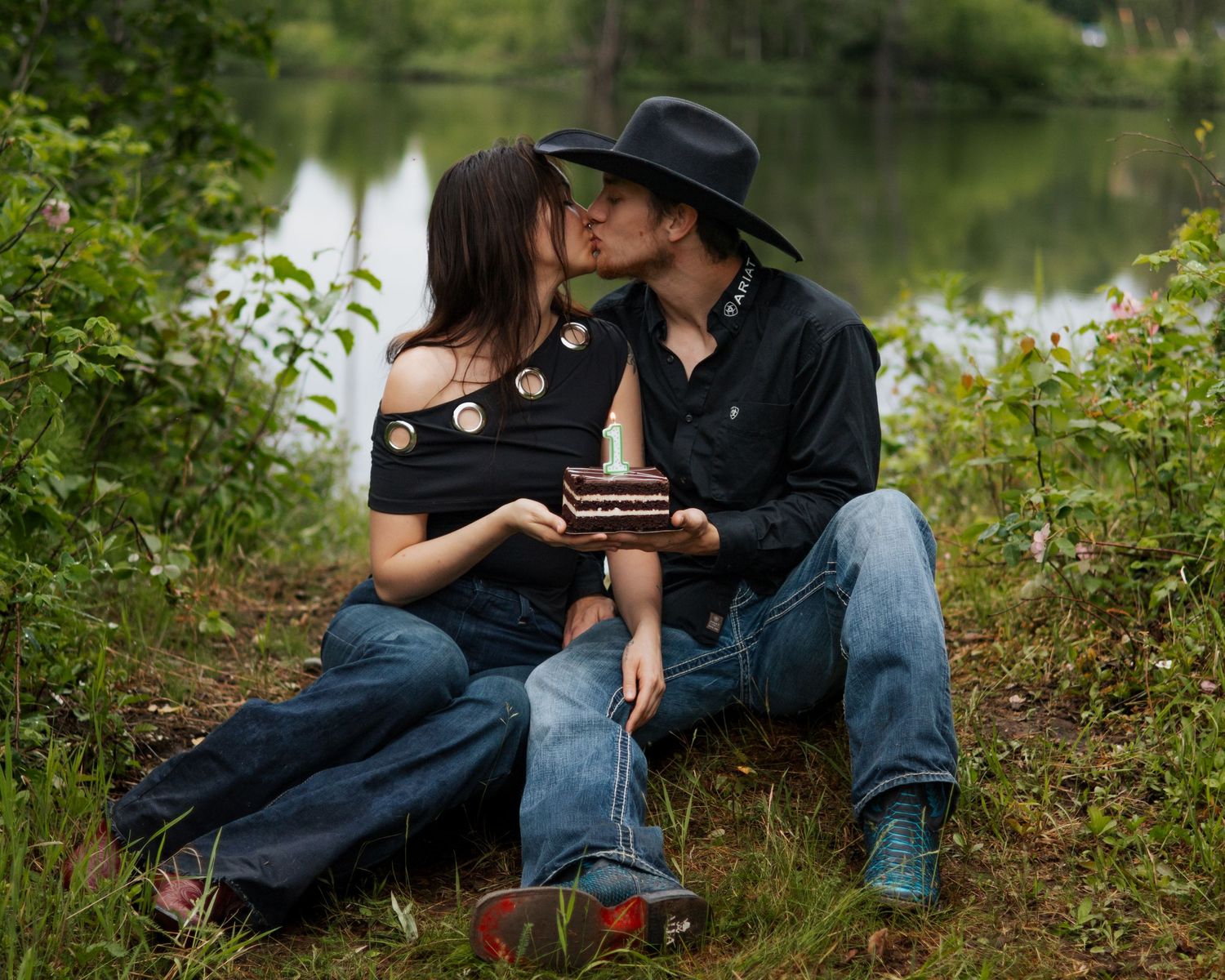 Couple kissing outdoors by a pond, man in black cowboy hat holding a small gift box, surrounded by lush green nature.