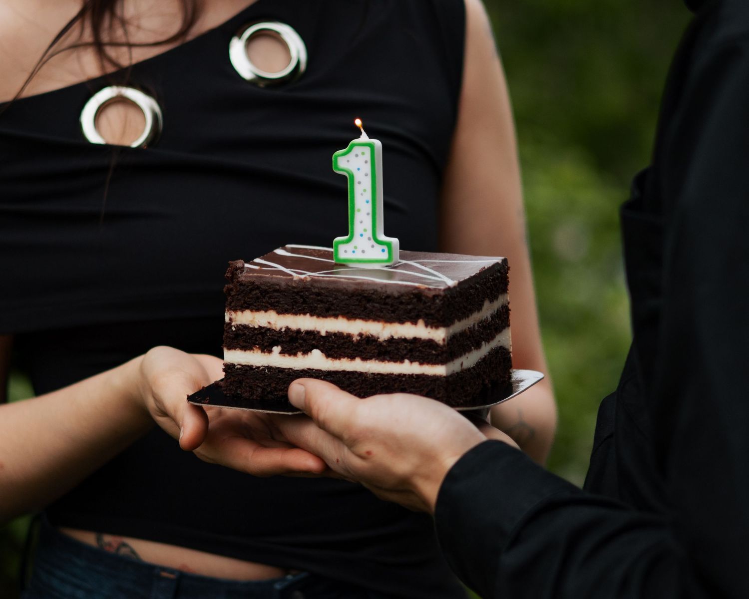 A chocolate layer cake with a number 1 candle on a plate, held by two people outdoors.