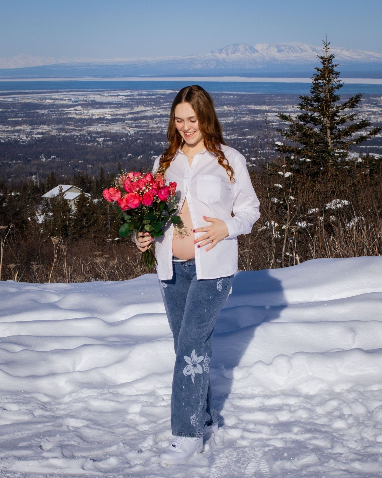Pregnant woman holding pink roses in snowy winter landscape with mountain view backdrop.