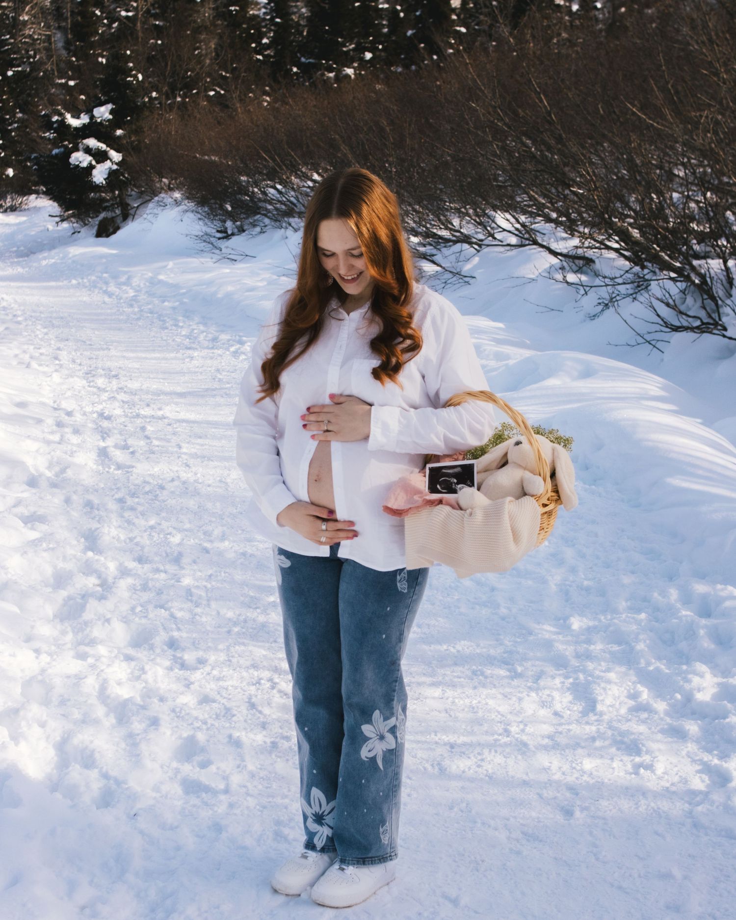Pregnant woman in white sweater and jeans holding ultrasound photo and basket in snowy winter landscape.