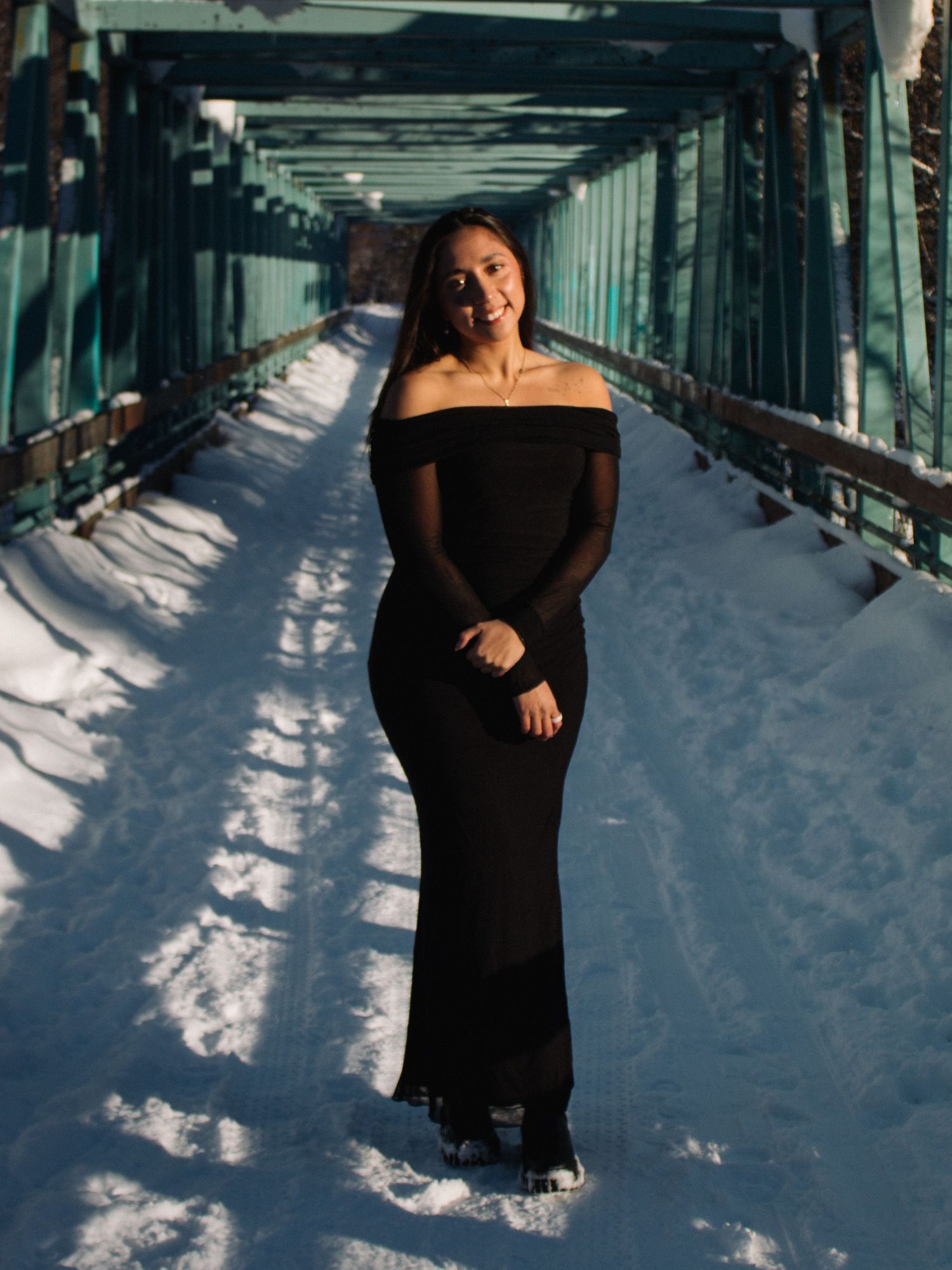 Woman in black off-shoulder gown poses on snowy bridge with teal metal railings in winter sunlight.