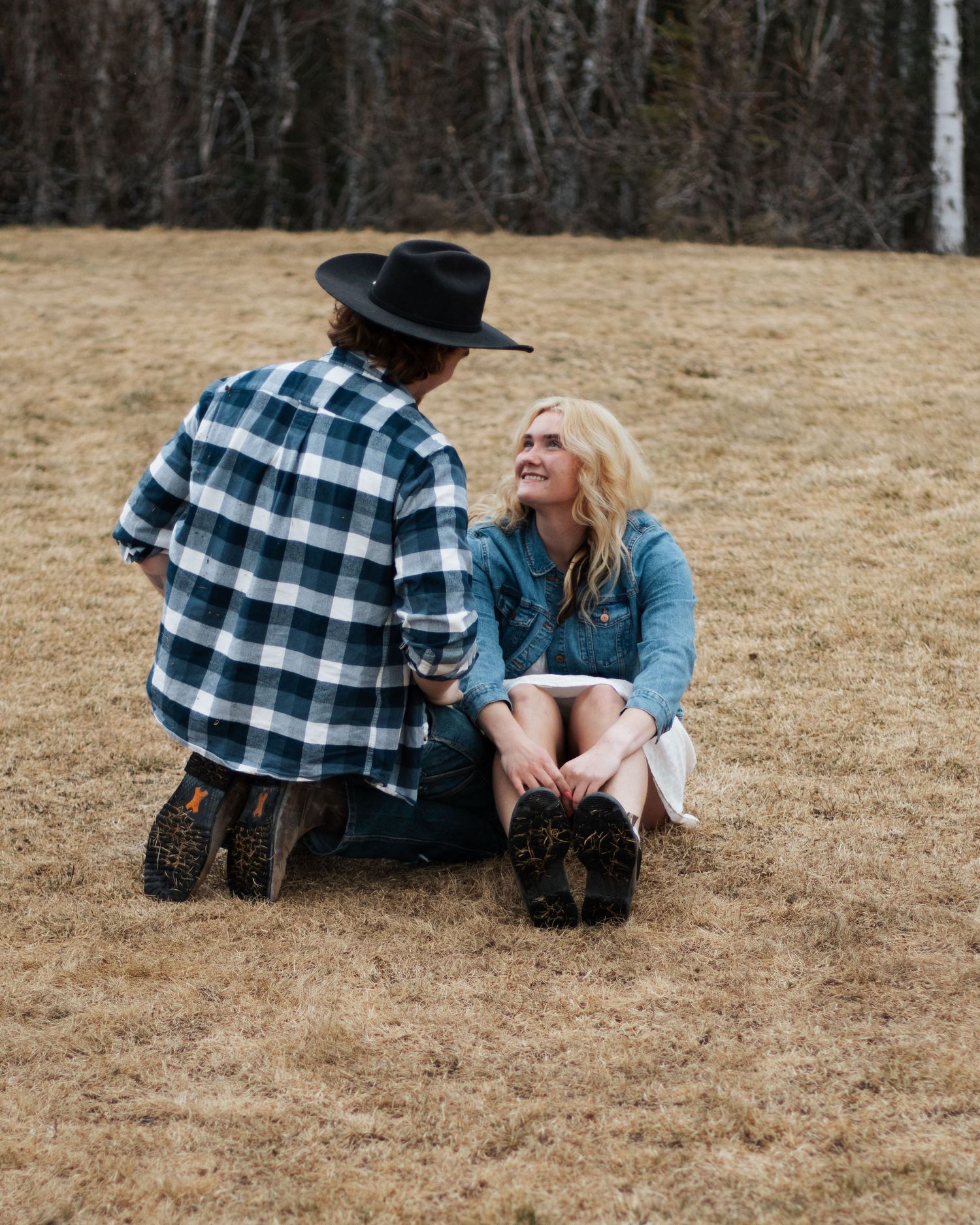 Two people sitting on dry grass in a field, one wearing a plaid shirt and cowboy hat, sharing a romantic moment outdoors.