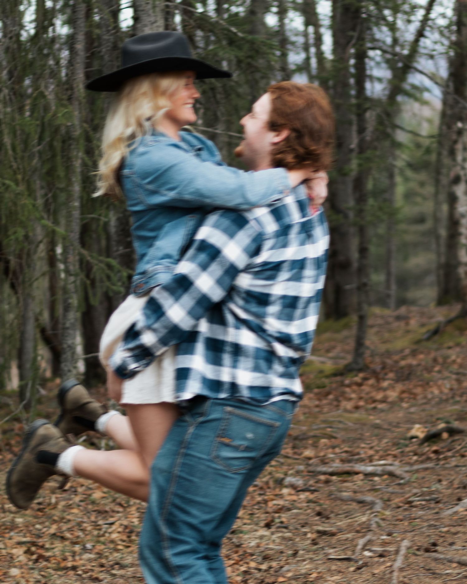 A man in a plaid shirt lifts a smiling blonde woman in a cowboy hat among forest trees.